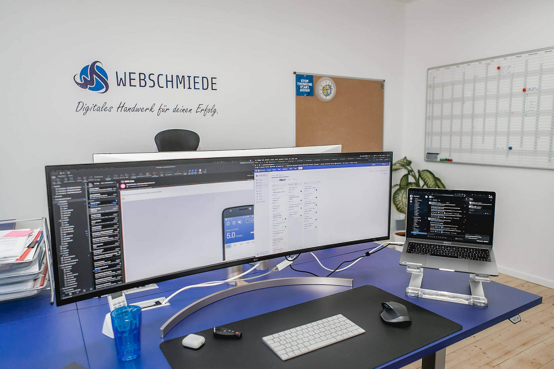 Modernes Büro mit großem Curved-Monitor, Laptop und Webschmiede Wandlogo für digitales Marketing und Webentwicklung.