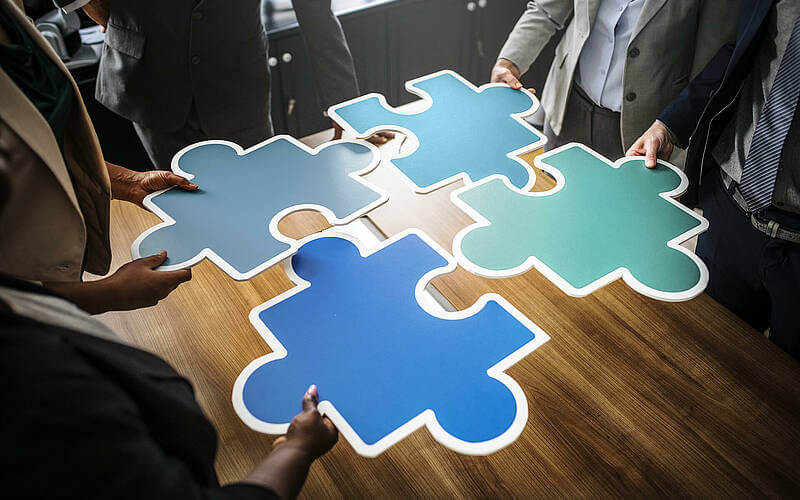 Menschen fügen große bunte Puzzleteile zusammen, symbolisieren Teamarbeit und Zusammenarbeit am Konferenztisch.