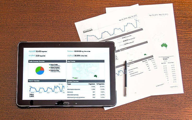 Tablet zeigt Website-Analyse-Dashboard mit Diagrammen und Statistiken, daneben gedruckte Berichte und ein Stift.