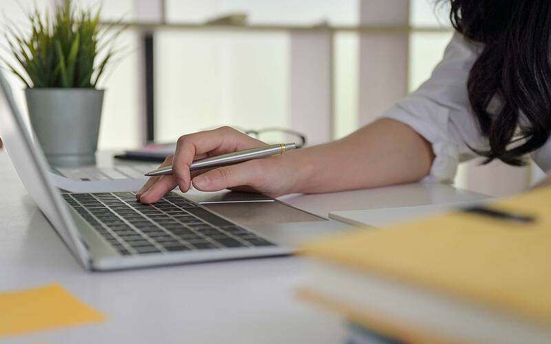 Person arbeitet am Laptop im Büro, hält einen Stift. Moderner Arbeitsplatz mit Pflanzen. Konzentration und Produktivität.