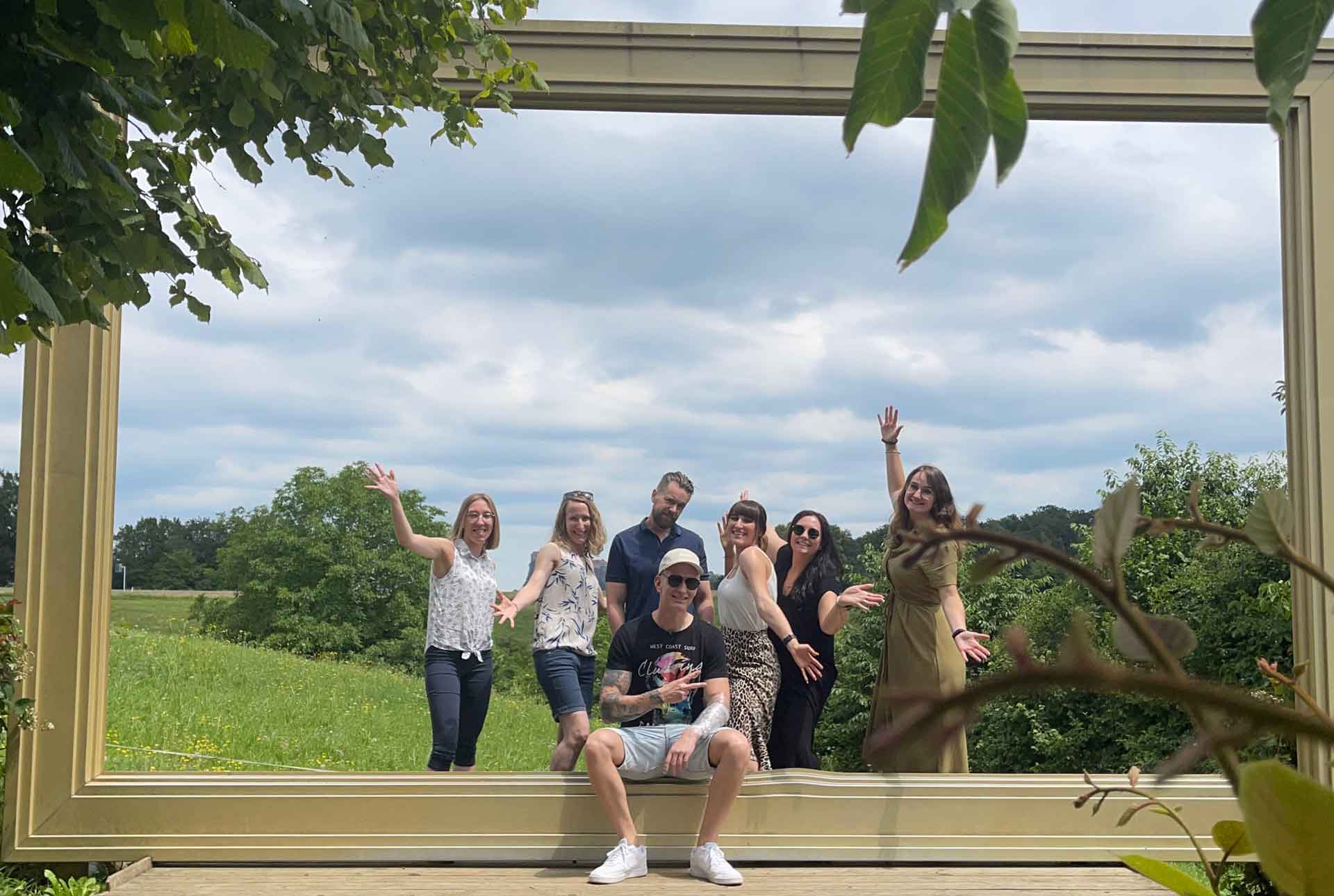 Gruppe von Menschen posiert fröhlich im Freien vor einem großen Bilderrahmen mit grüner Landschaft im Hintergrund.