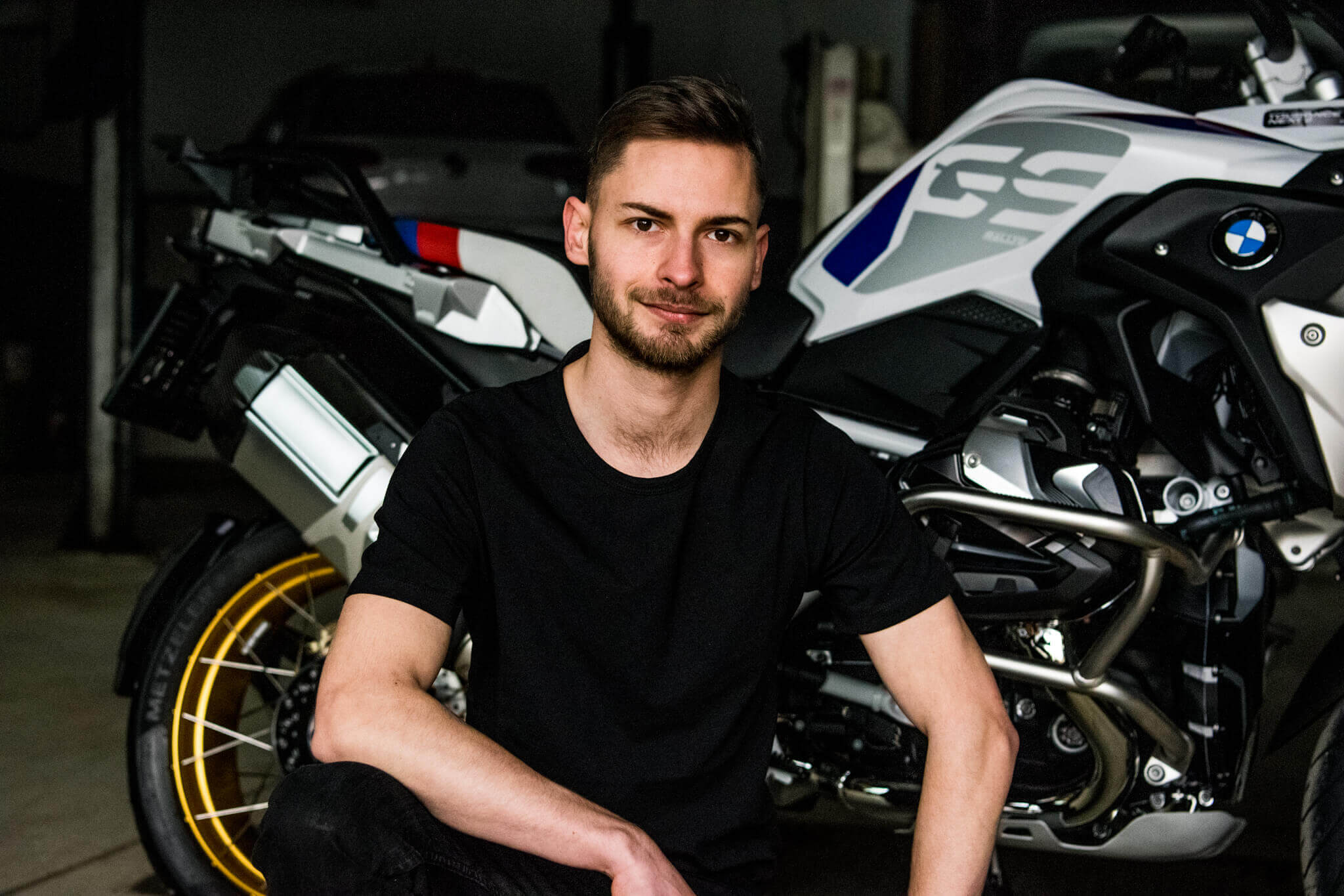 Mann in schwarzem T-Shirt posiert neben einem BMW Motorrad in einer Garage.