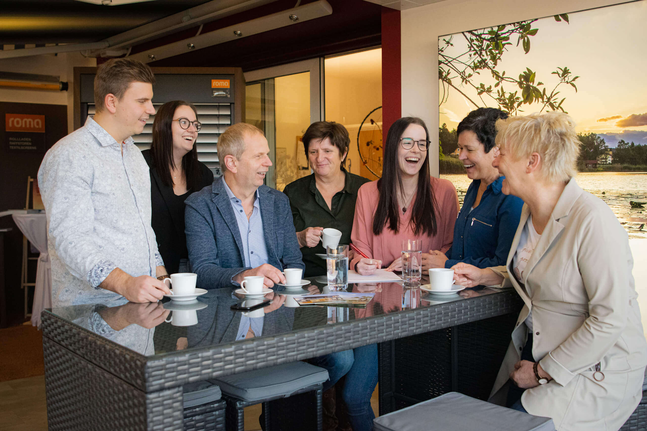 Gruppe von Menschen bei einem Meeting, lachend und Kaffee trinkend, in modernem Büro mit Naturbild im Hintergrund.