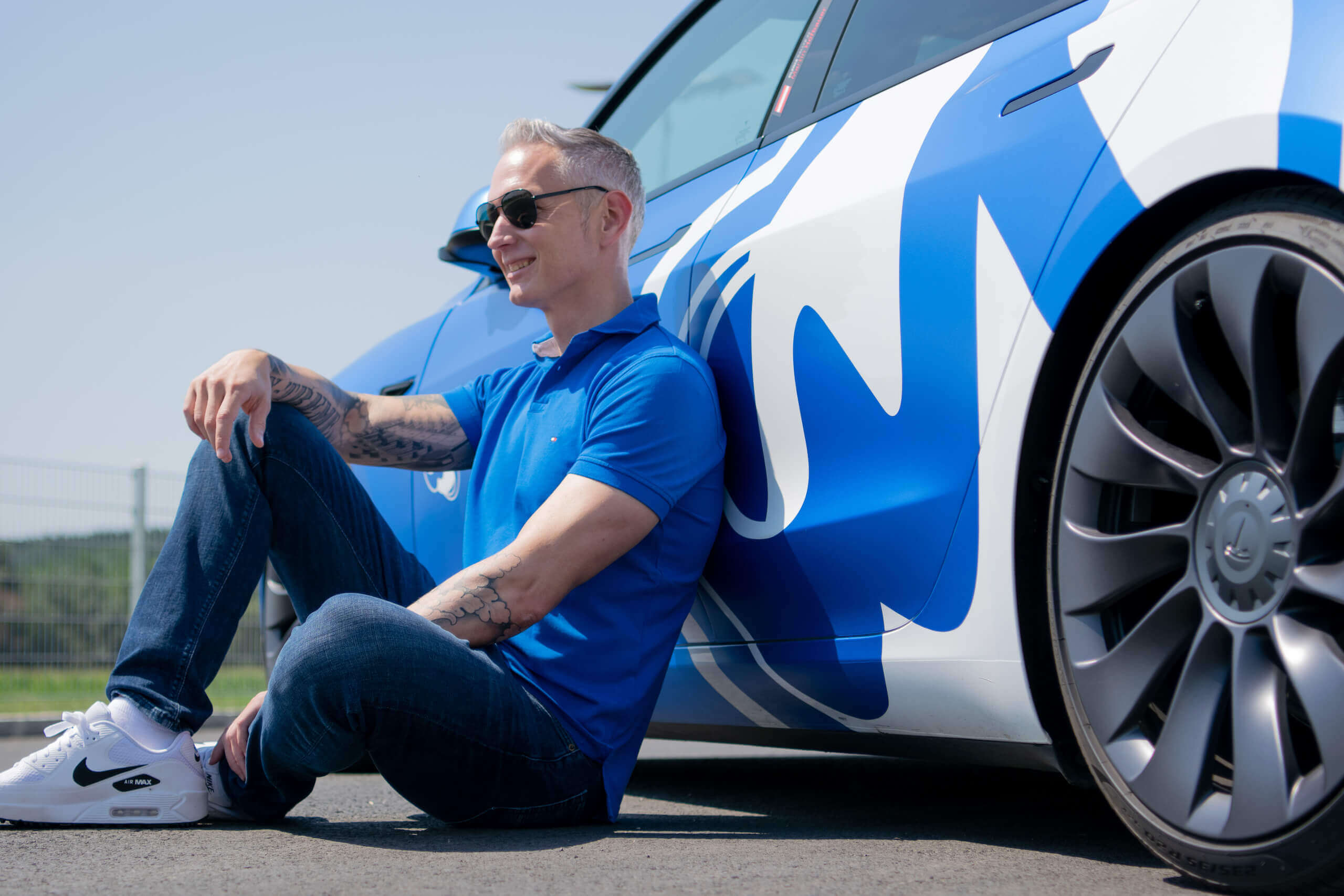 Mann sitzt entspannt neben blau-weißem Auto auf Straße, trägt Sonnenbrille und Jeans, sonniger Tag.