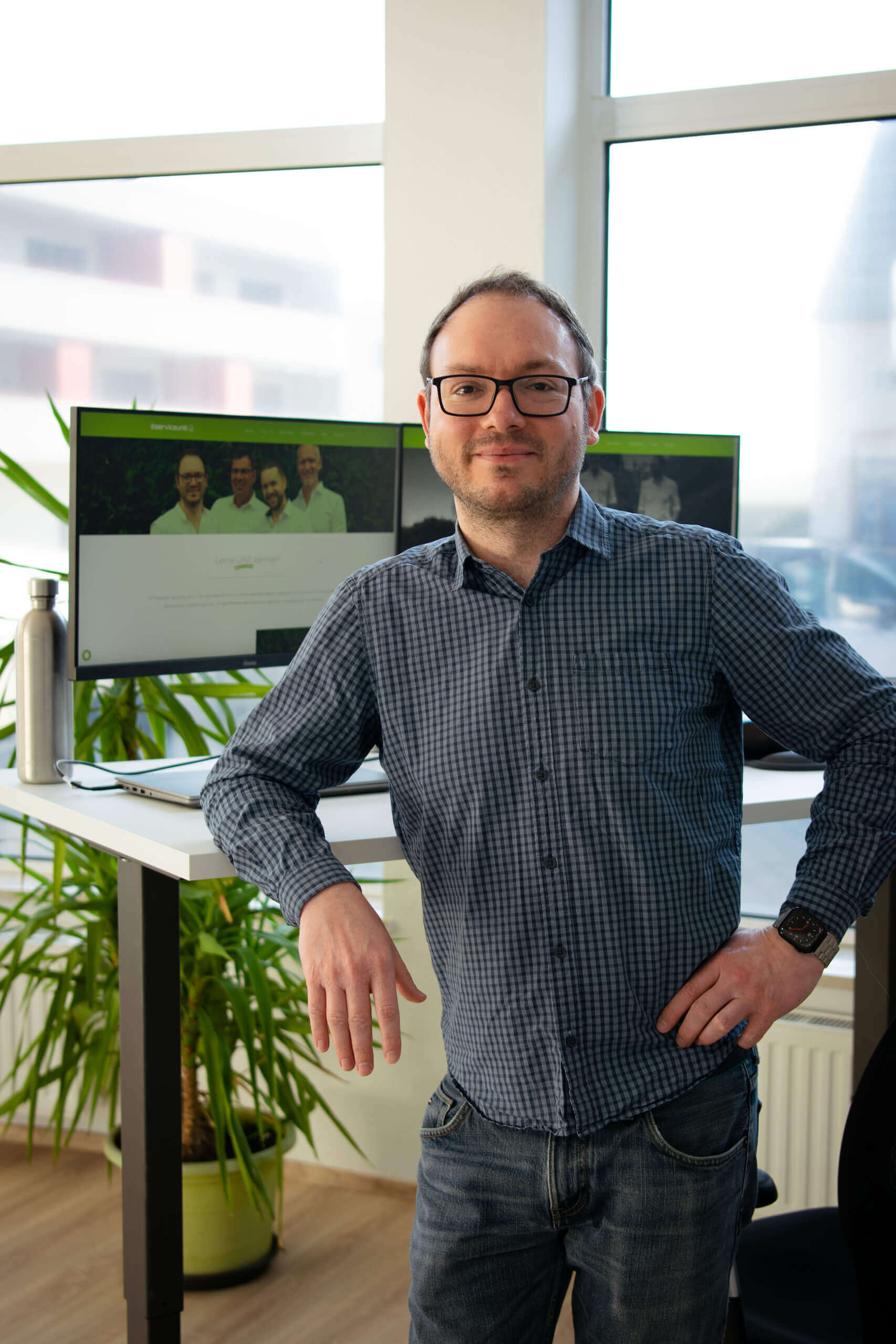 Mann im Büro stehend vor Schreibtisch mit Monitor und Pflanze im Hintergrund, lächelnd in die Kamera.