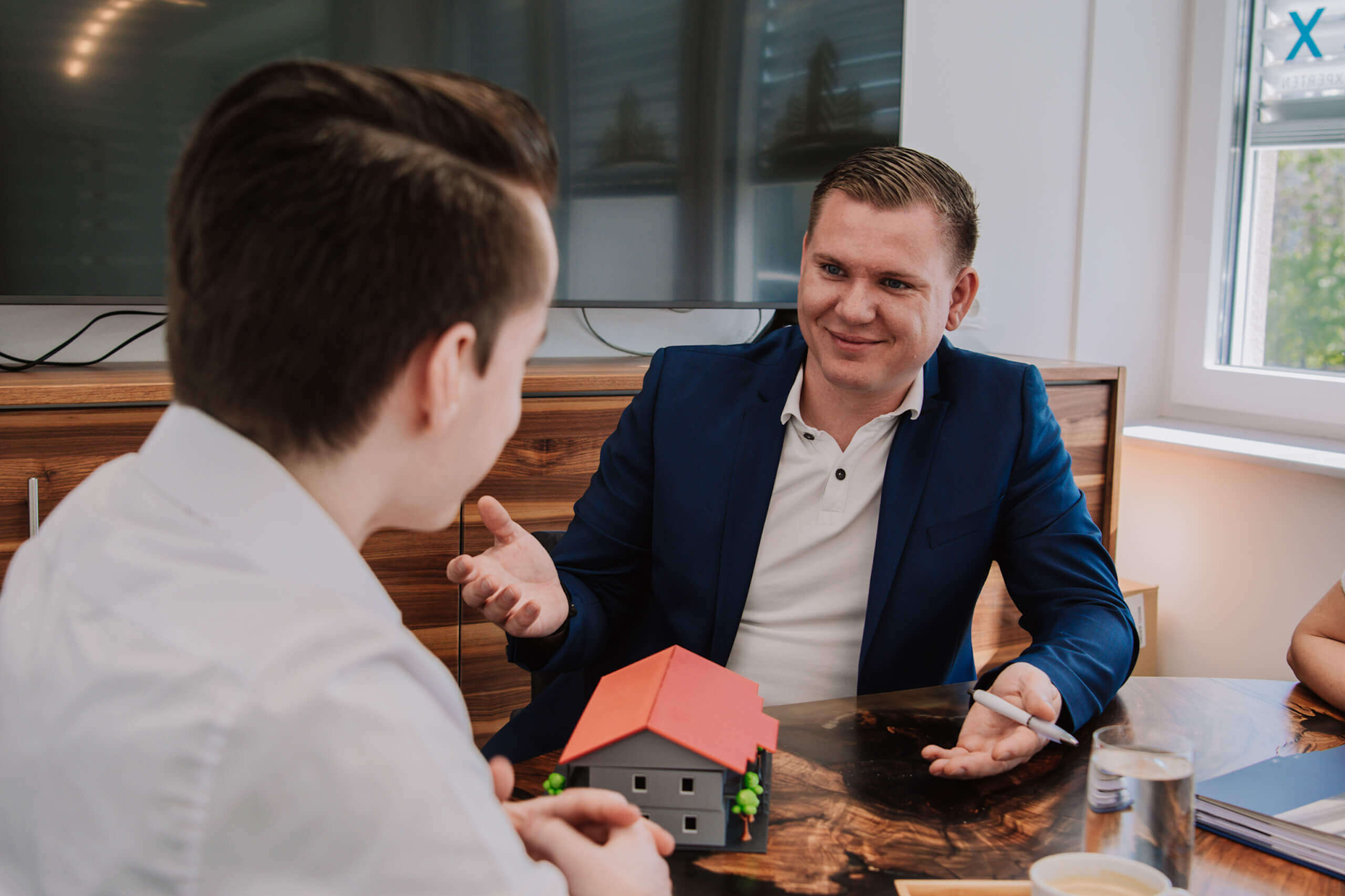 Beratungsgespräch für Immobilienkauf, zwei Männer diskutieren über ein Modellhaus am Konferenztisch.