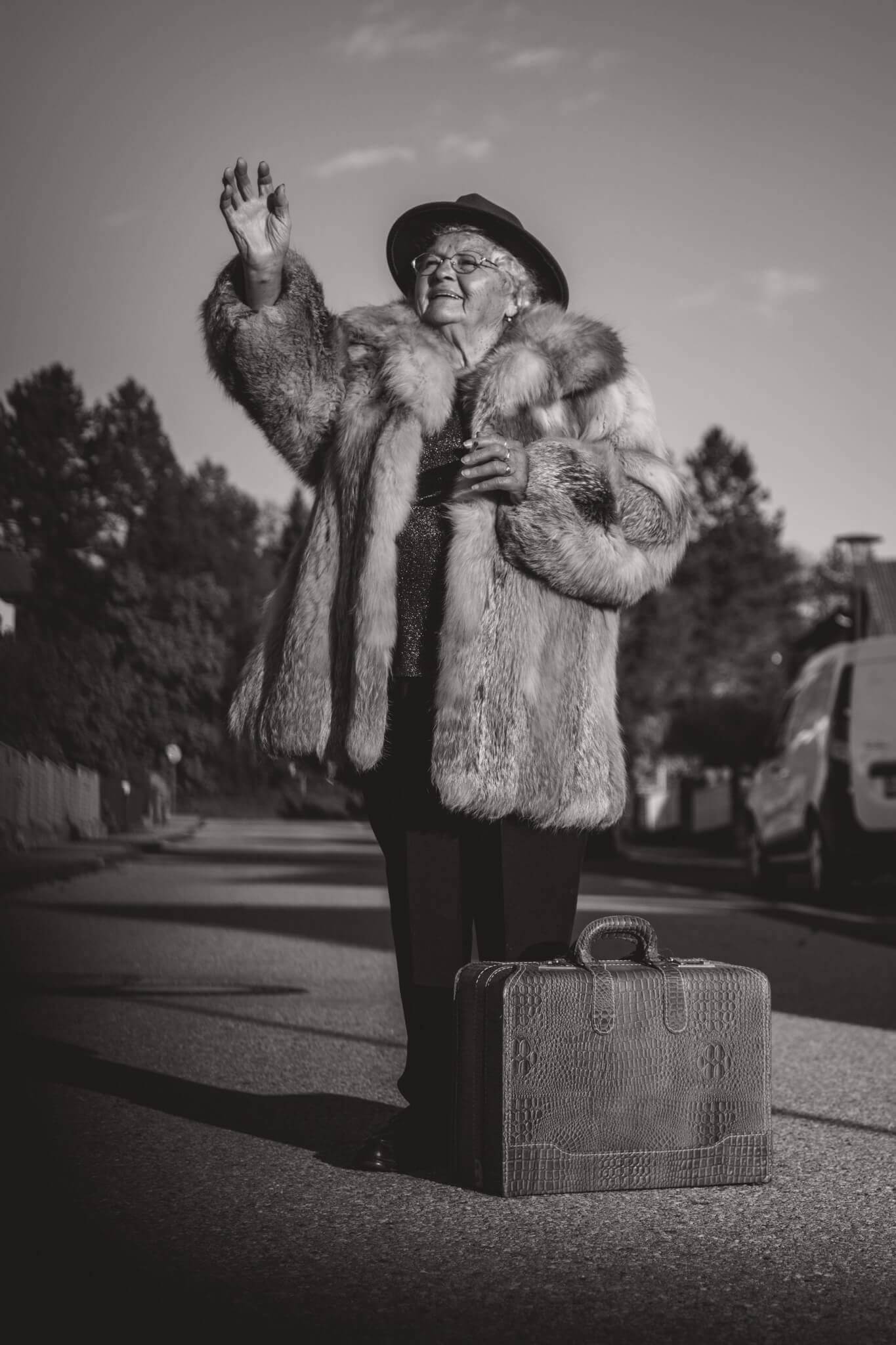 Ältere Frau in Pelzjacke winkt auf Straße, vintage Reisekoffer, nostalgische Reiseatmosphäre, Schwarz-Weiß-Fotografie.