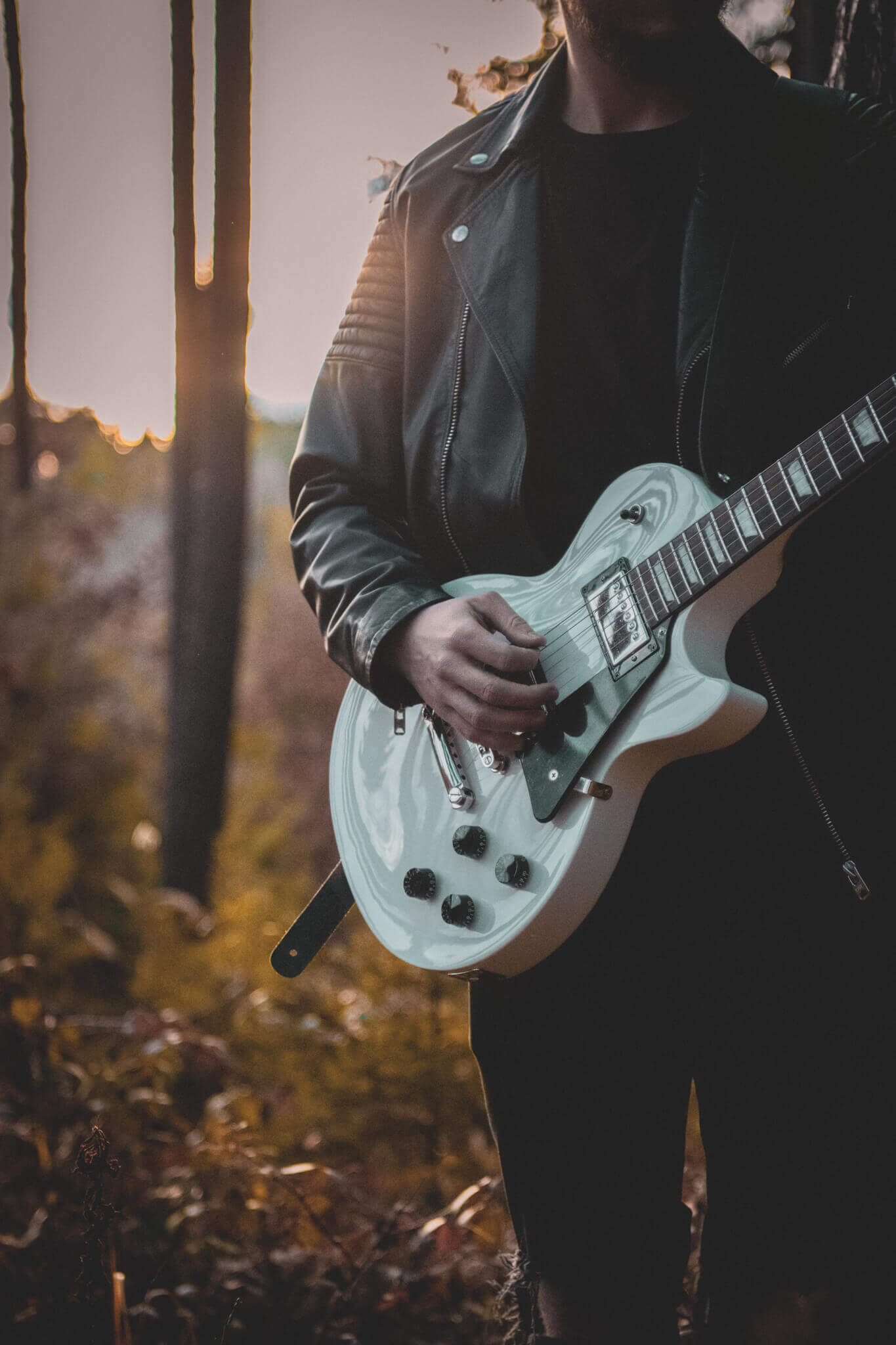 Gitarrist in Lederjacke spielt weiße E-Gitarre bei Sonnenuntergang im Wald. Atmosphärische Musikaufnahme draußen.