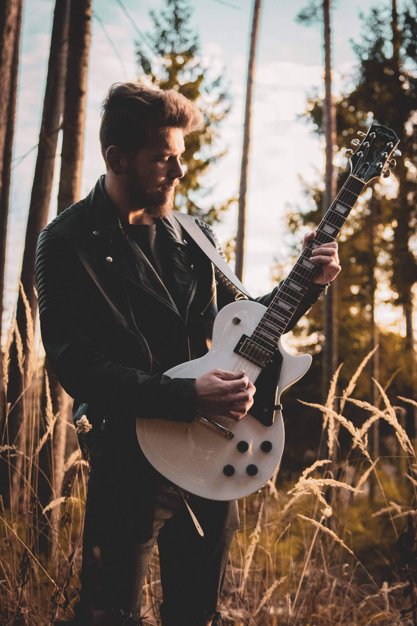 Mann spielt E-Gitarre im Sonnenuntergang, umgeben von Bäumen und hohen Gräsern, in Lederjacke.