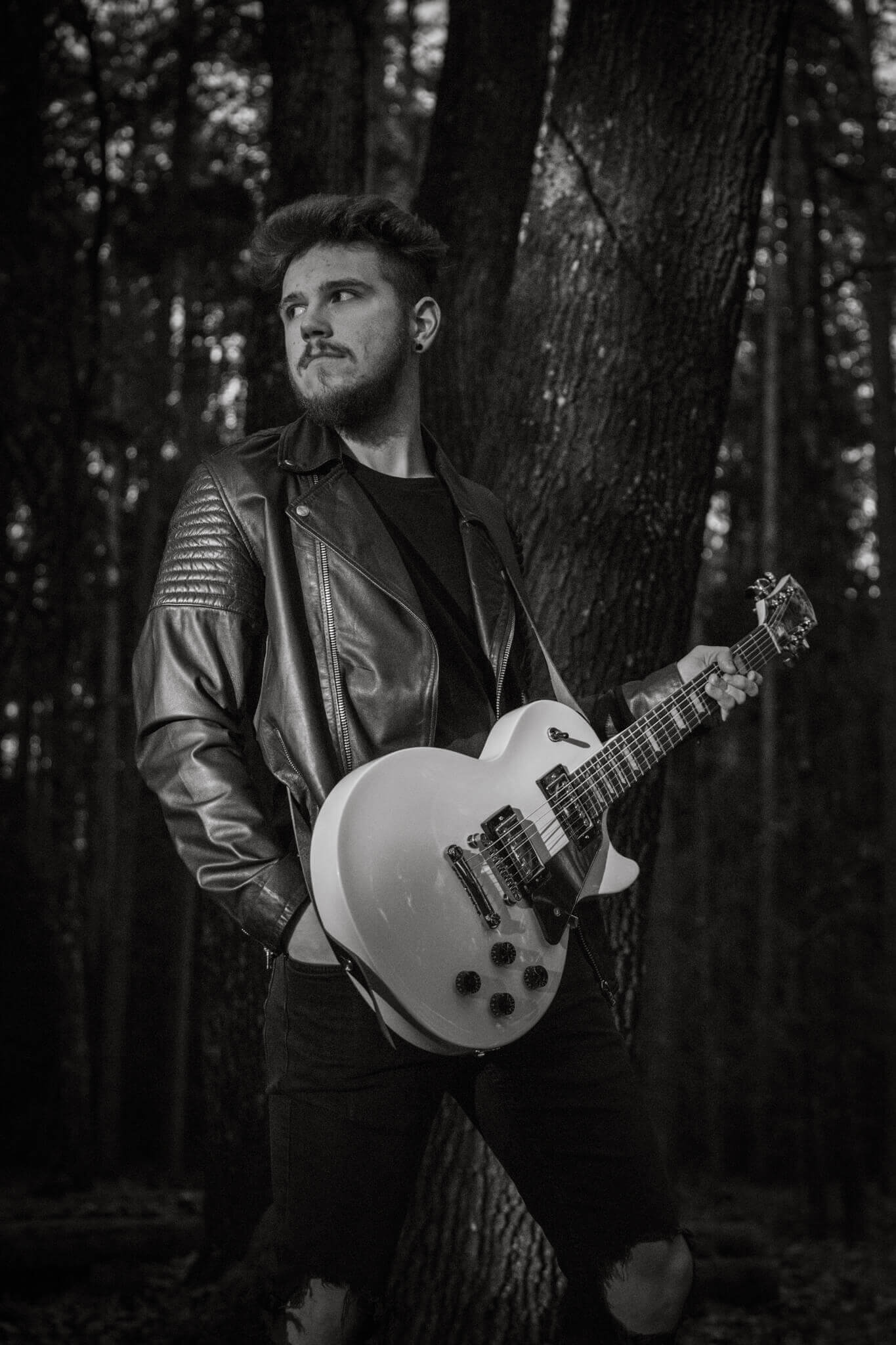 Musiker im Wald mit E-Gitarre und Lederjacke, schwarz-weiß Foto, Rock-Thema, stimmungsvolles Portrait.