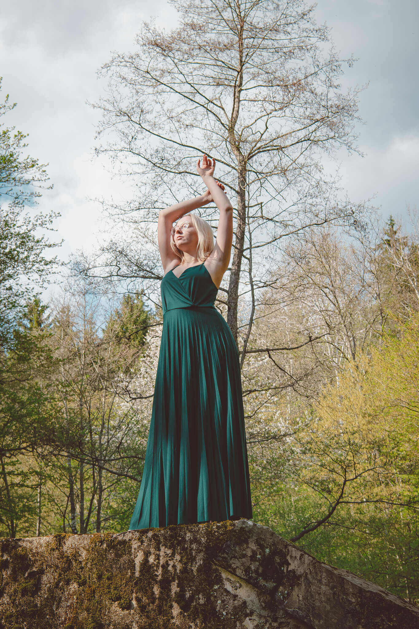 Frau im langen grünen Kleid posiert auf Felsen im Wald, umgeben von Bäumen und blauem Himmel im Hintergrund.