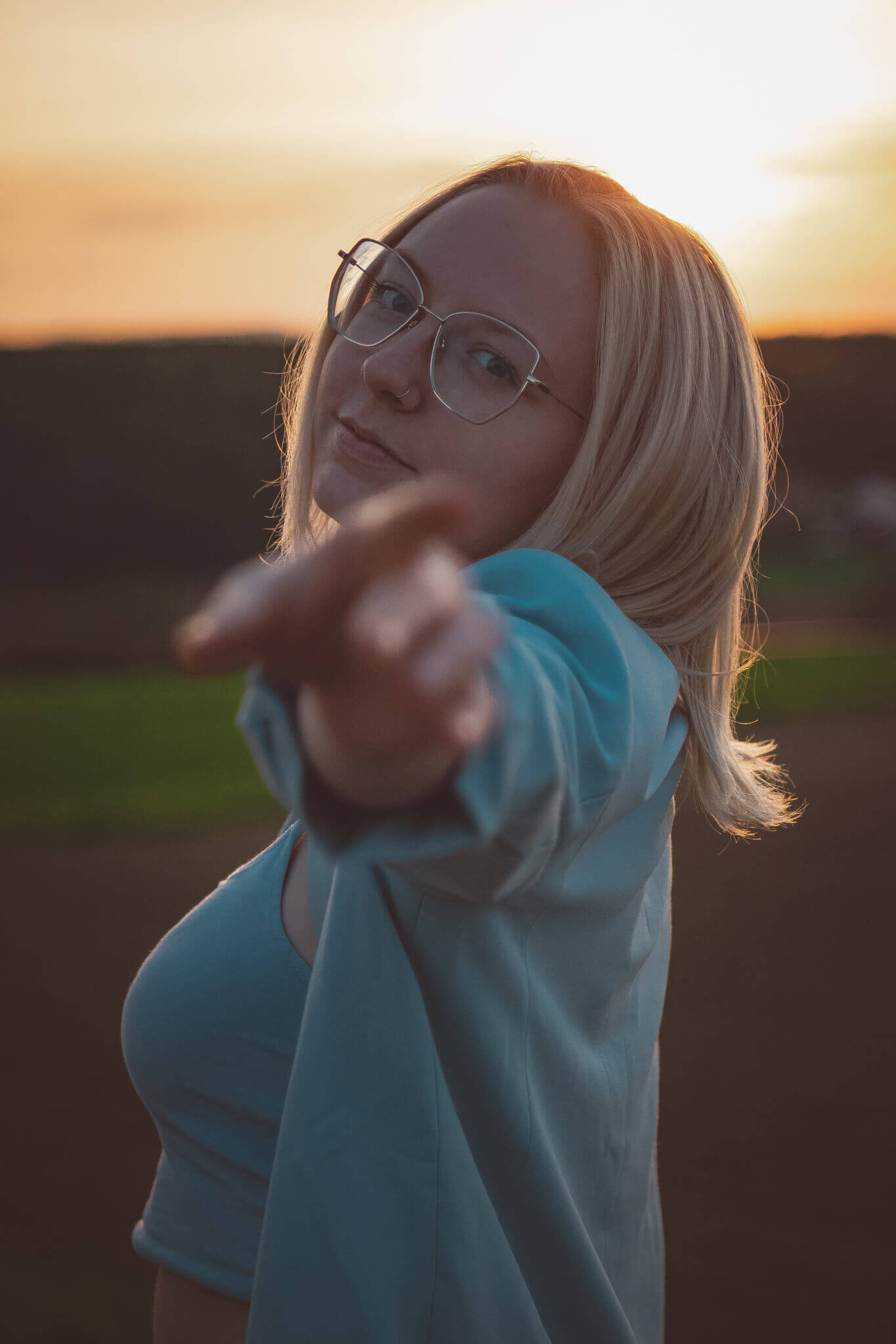 Person zeigt auf die Kamera bei Sonnenuntergang, trägt Brille und hellblauen Blazer, mit inspirierendem Blick.