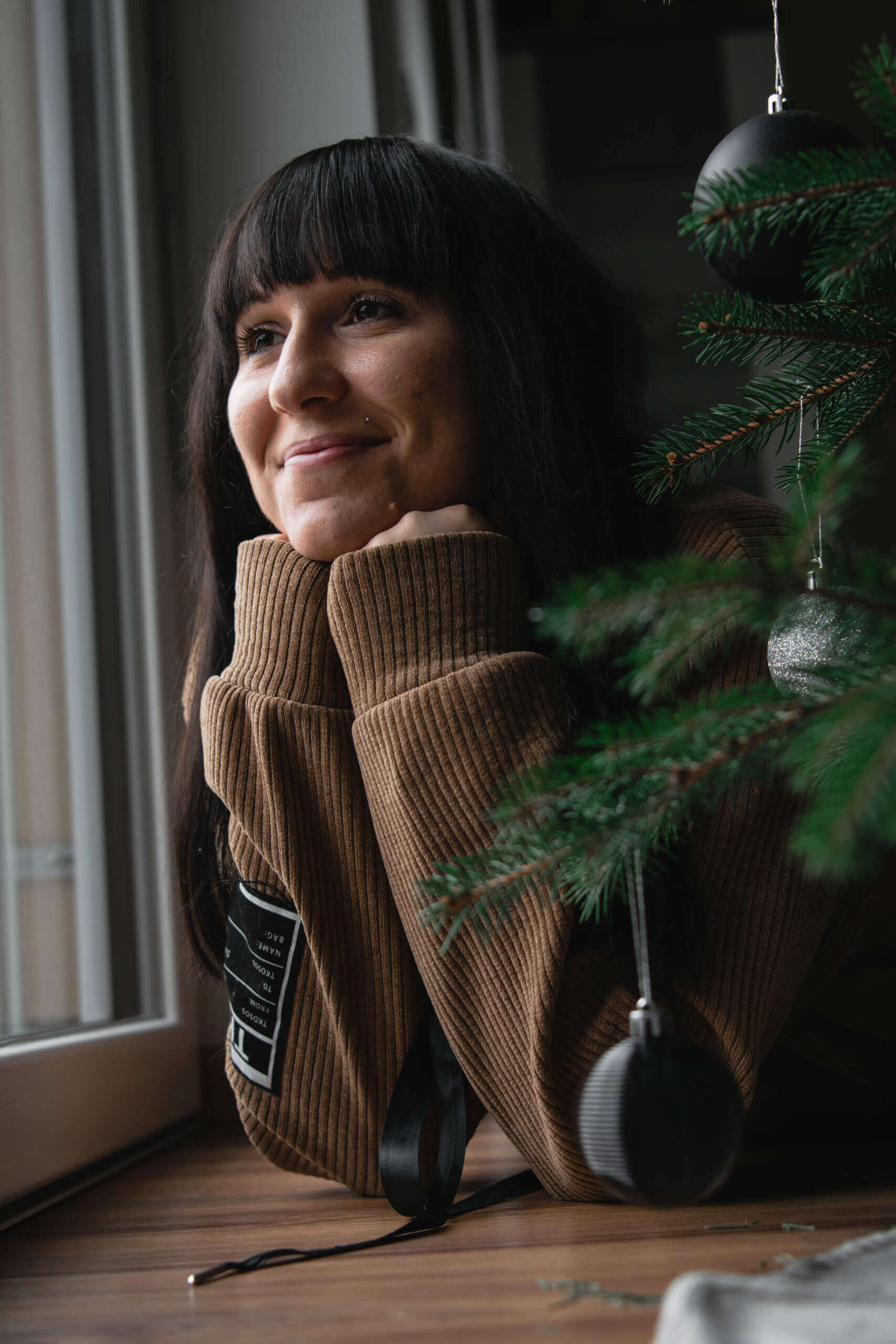 Frau in braunem Pullover lächelt nachdenklich am Fenster mit Weihnachtsbaum im Vordergrund.