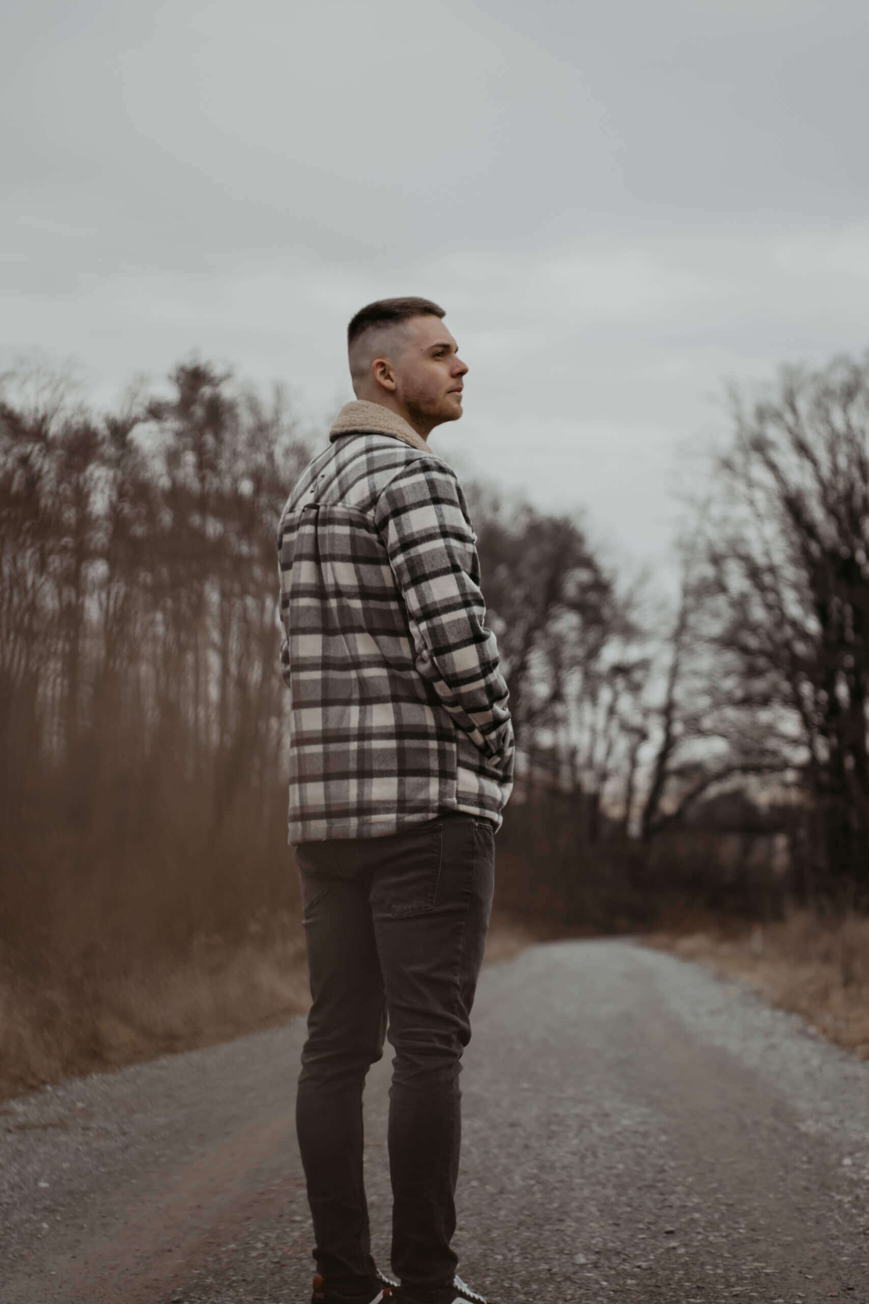 Mann in karierter Jacke steht auf ländlichem Weg, Bäume im Hintergrund, bewölkter Himmel.