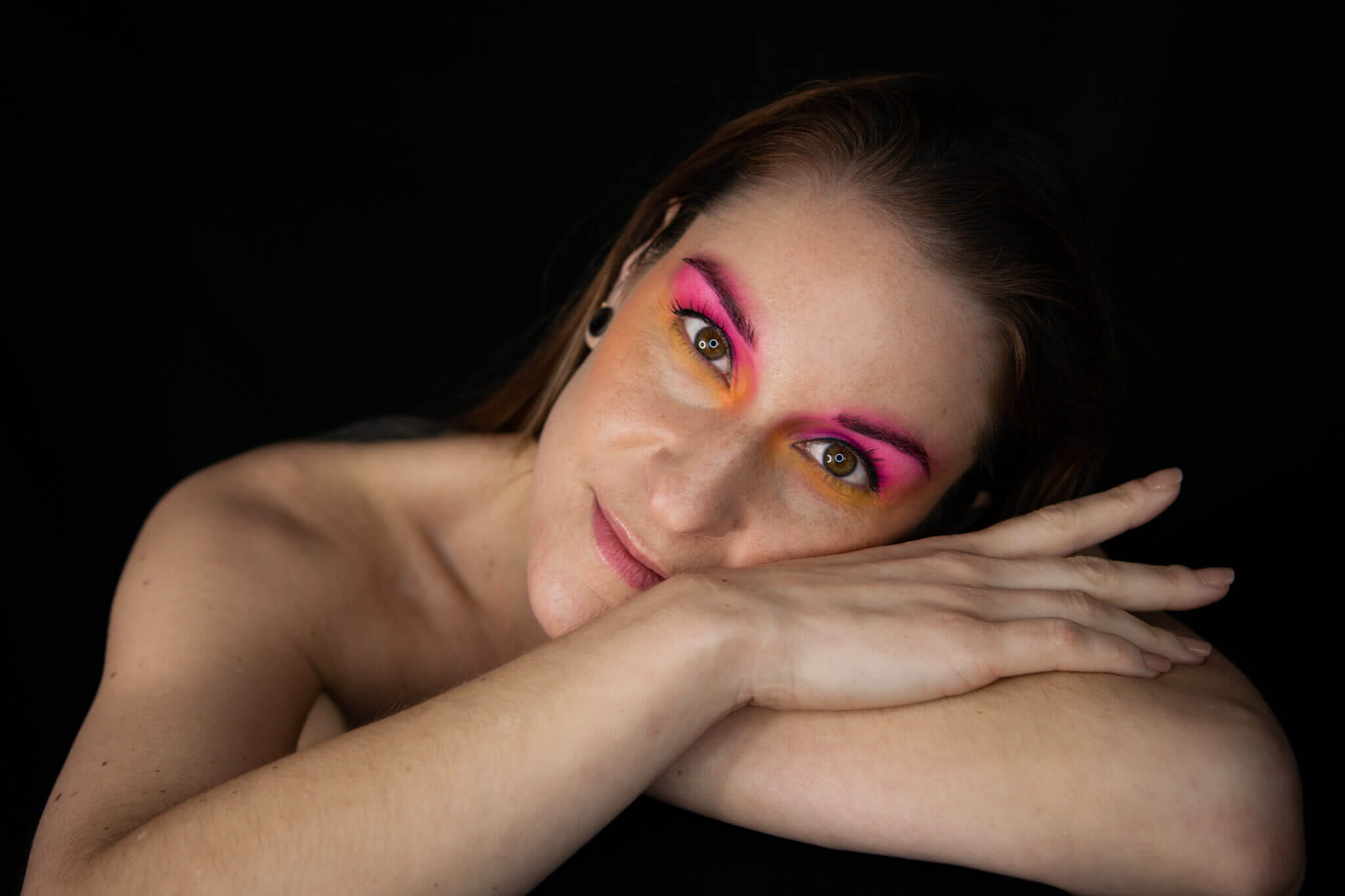 Frau mit kreativem Make-up in Pink und Orange lehnt den Kopf auf ihre Arme und blickt in die Kamera.