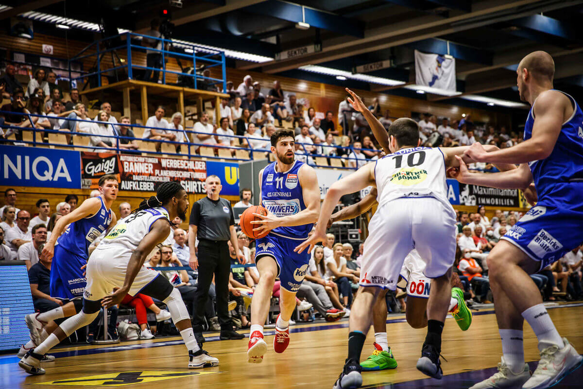 Basketballspiel in einer vollen Halle, Spieler in Aktion beim Dribbeln, umgeben von verteidigenden Gegnern.