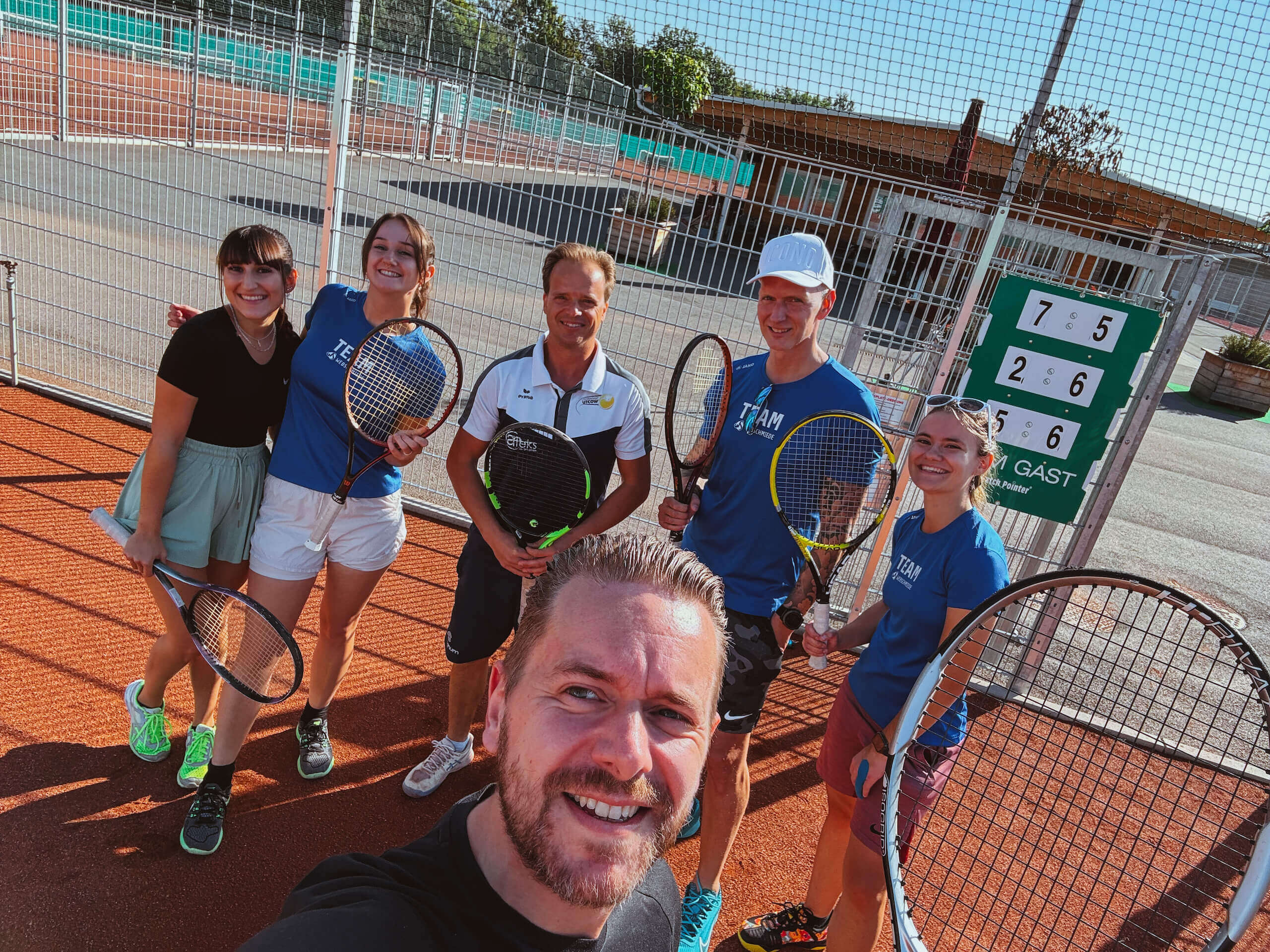 Tennisgruppe beim Spiel auf Sandplatz, fröhlich posierend mit Schlägern, im Hintergrund Anzeigetafel mit Punktestand.