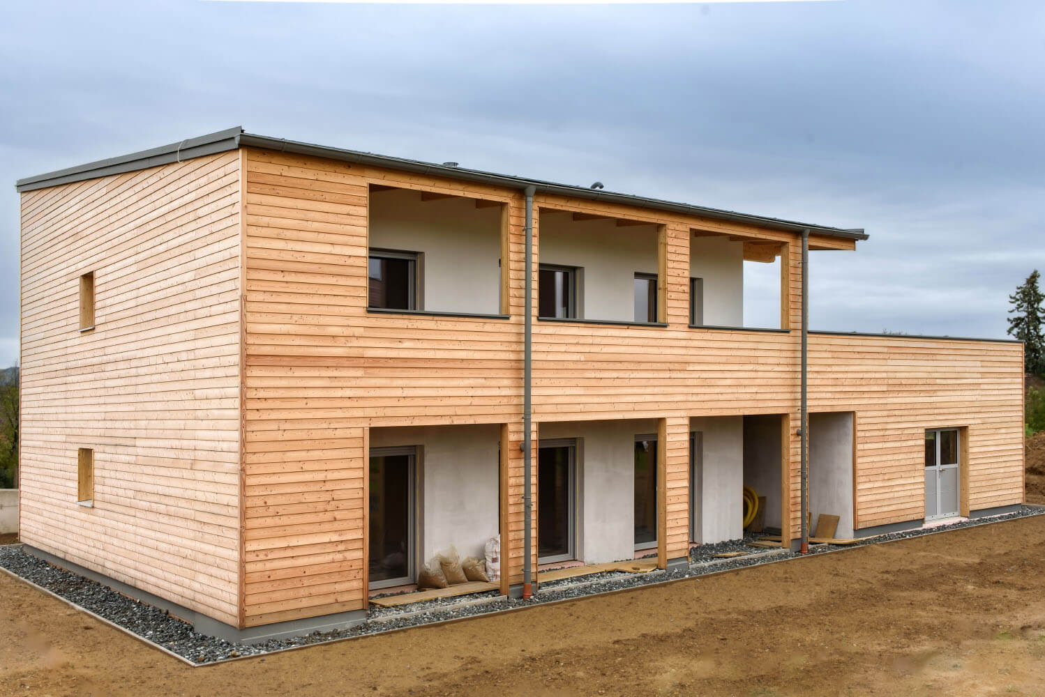 Moderner Holzbau: Zweistöckiges Gebäude mit Holzverkleidung auf offenem Grundstück vor bedecktem Himmel.