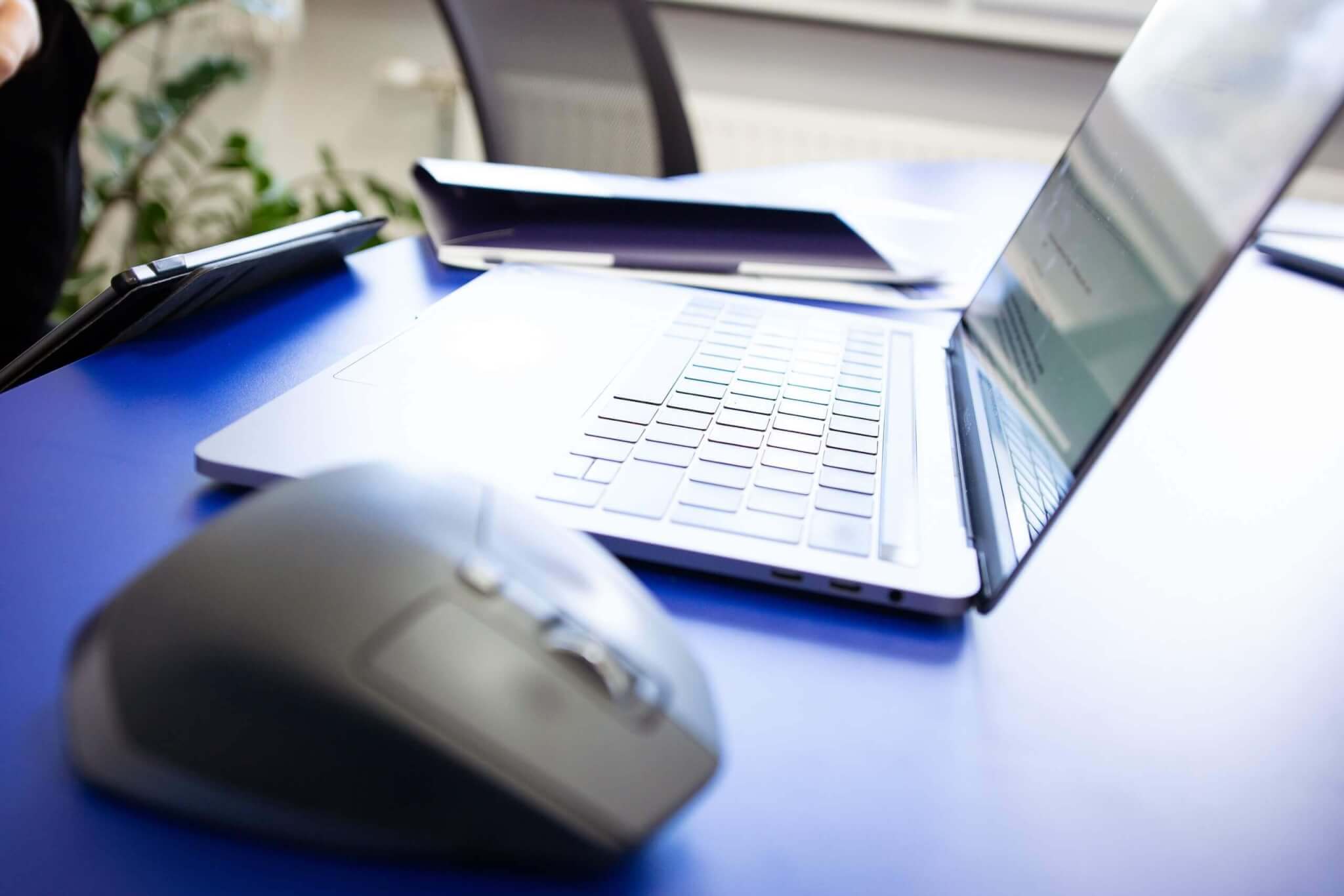 Laptop und Maus auf blauem Schreibtisch für modernes Arbeiten im Büro oder Homeoffice.