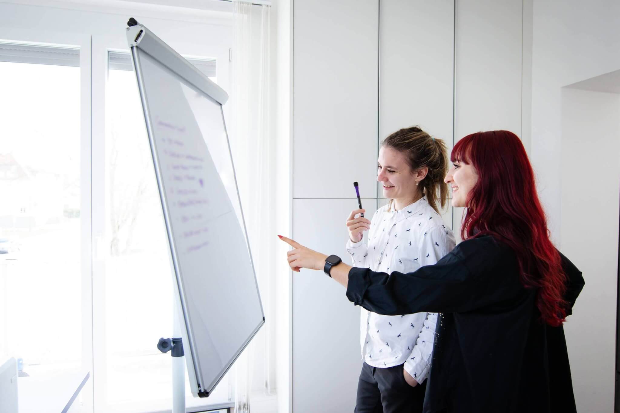 Zwei Frauen besprechen Notizen auf einem Whiteboard im Büro, lächelnd und fokussiert auf die Zusammenarbeit.