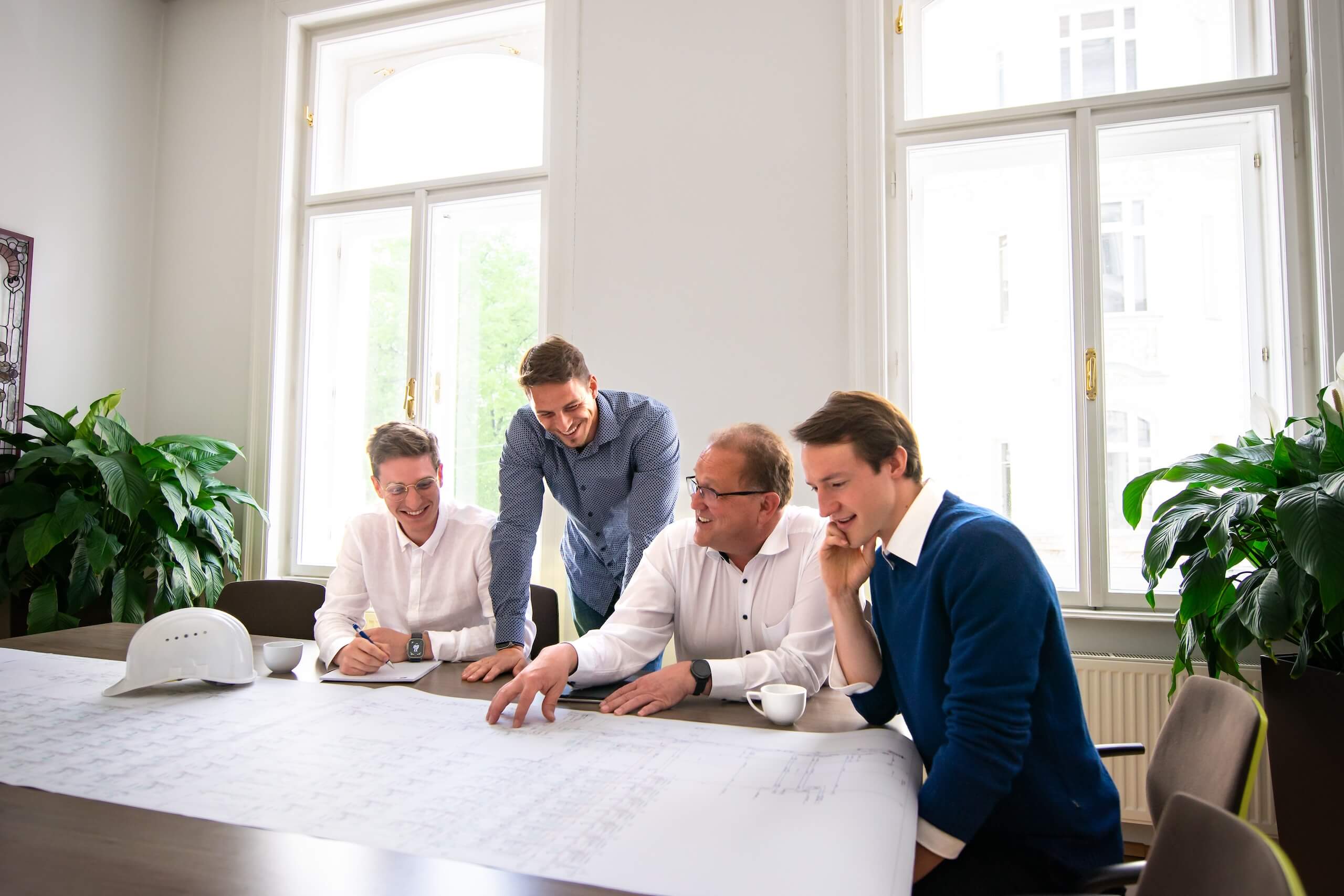 Gruppe von Geschäftsleuten bei einer Besprechung am Konferenztisch mit Bauplänen und Pflanzen im Hintergrund.