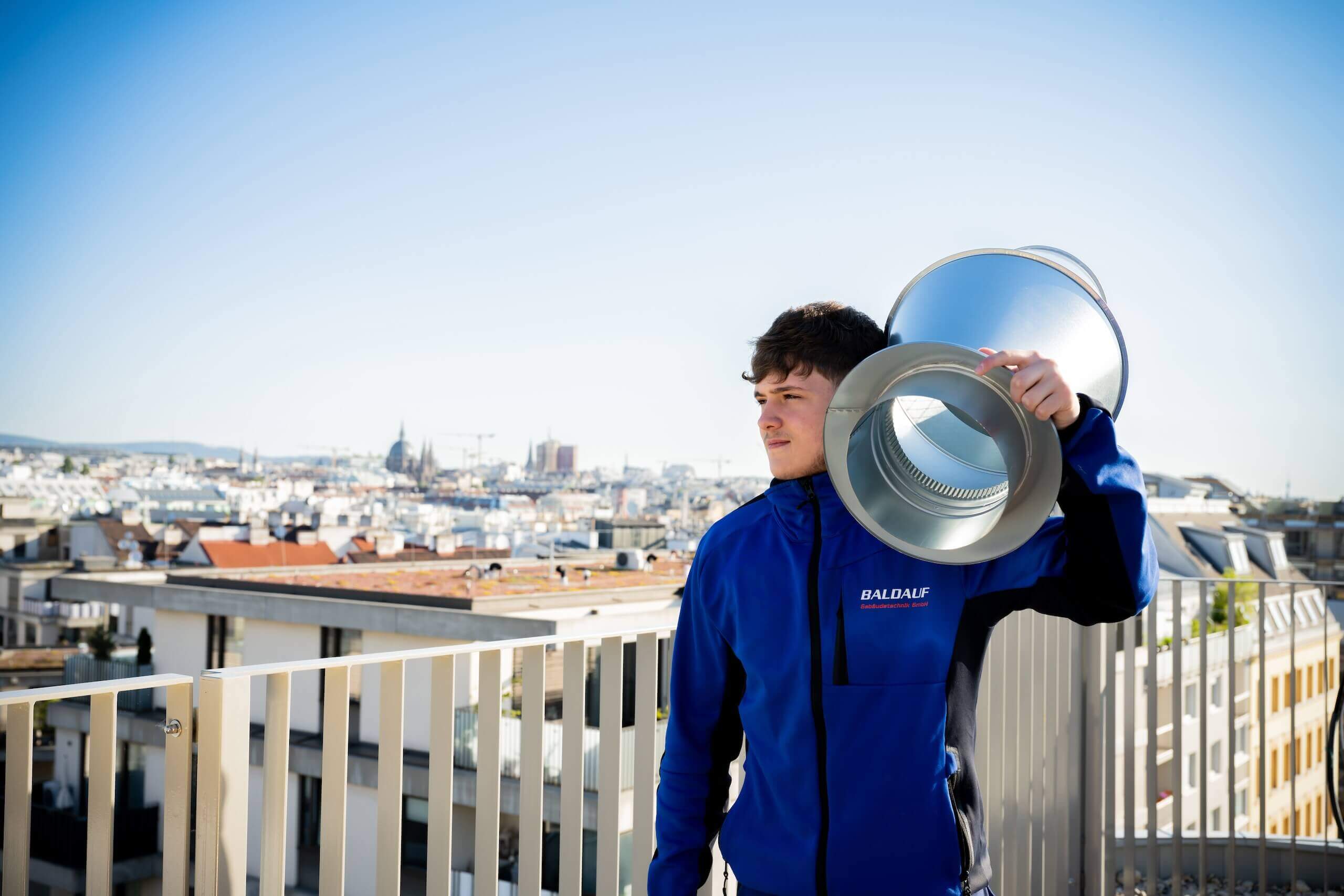 Mann im blauen Arbeitsanzug mit Metallventil auf Dach in urbaner Umgebung bei klarem Himmel.