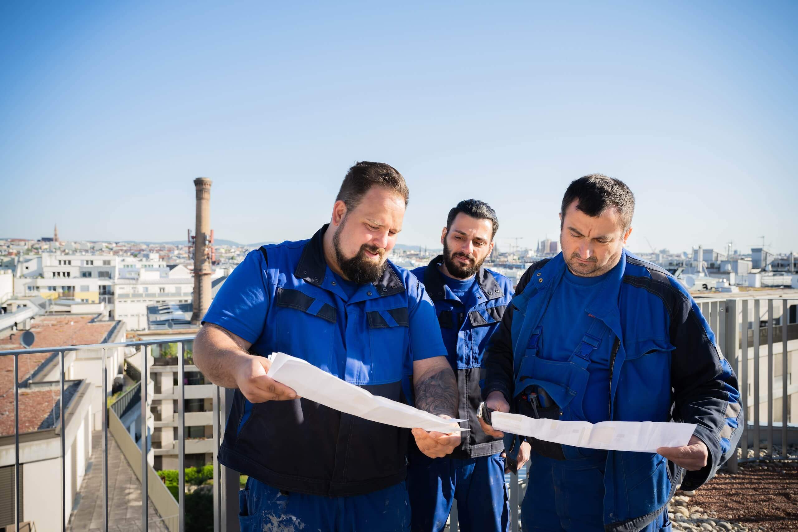 Drei Bauarbeiter in blauer Arbeitskleidung begutachten Baupläne auf einer Dachterrasse in der Stadt.