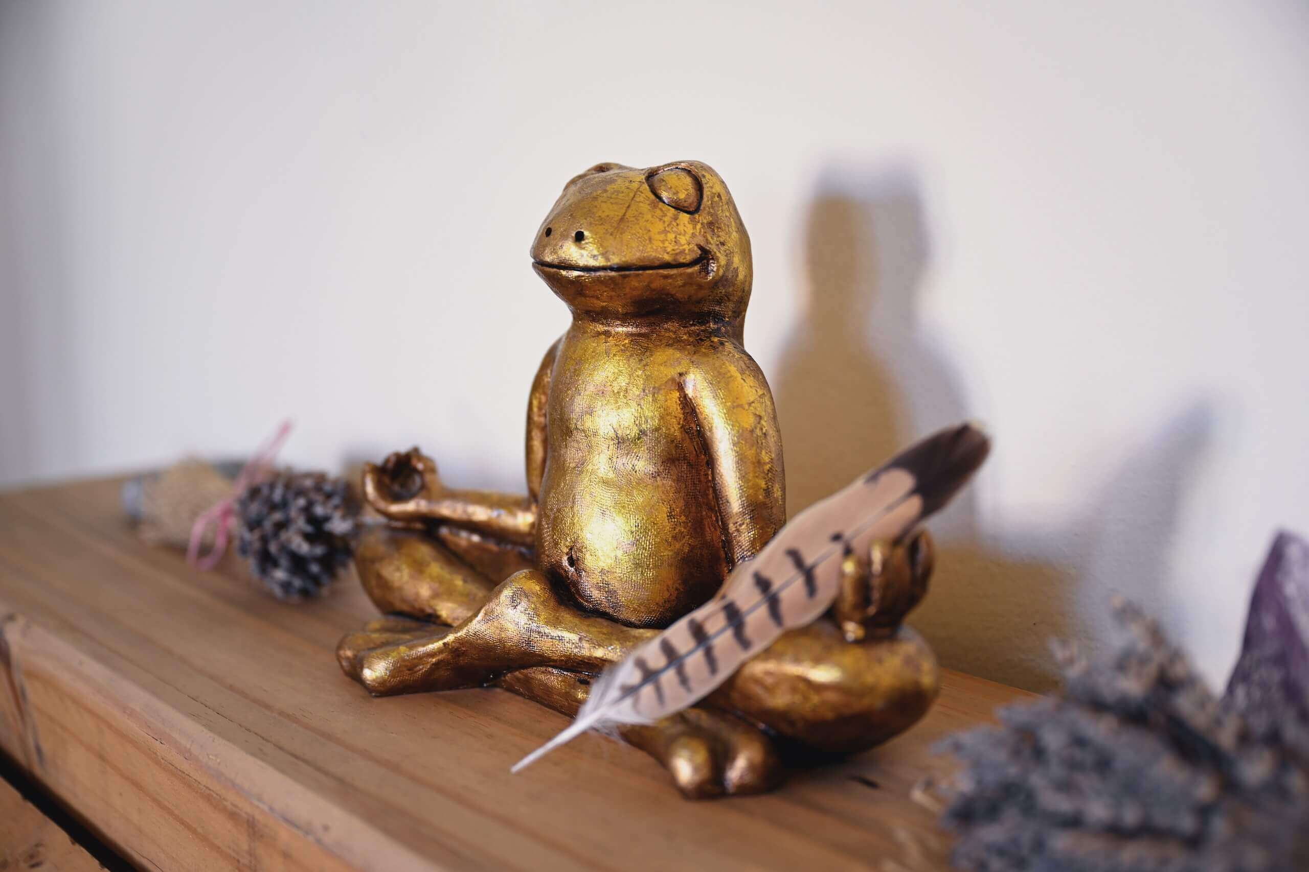 Goldene Froschstatue im Meditationssitz mit Feder auf Holzregal, umgeben von Naturmaterialien, friedliche Atmosphäre.