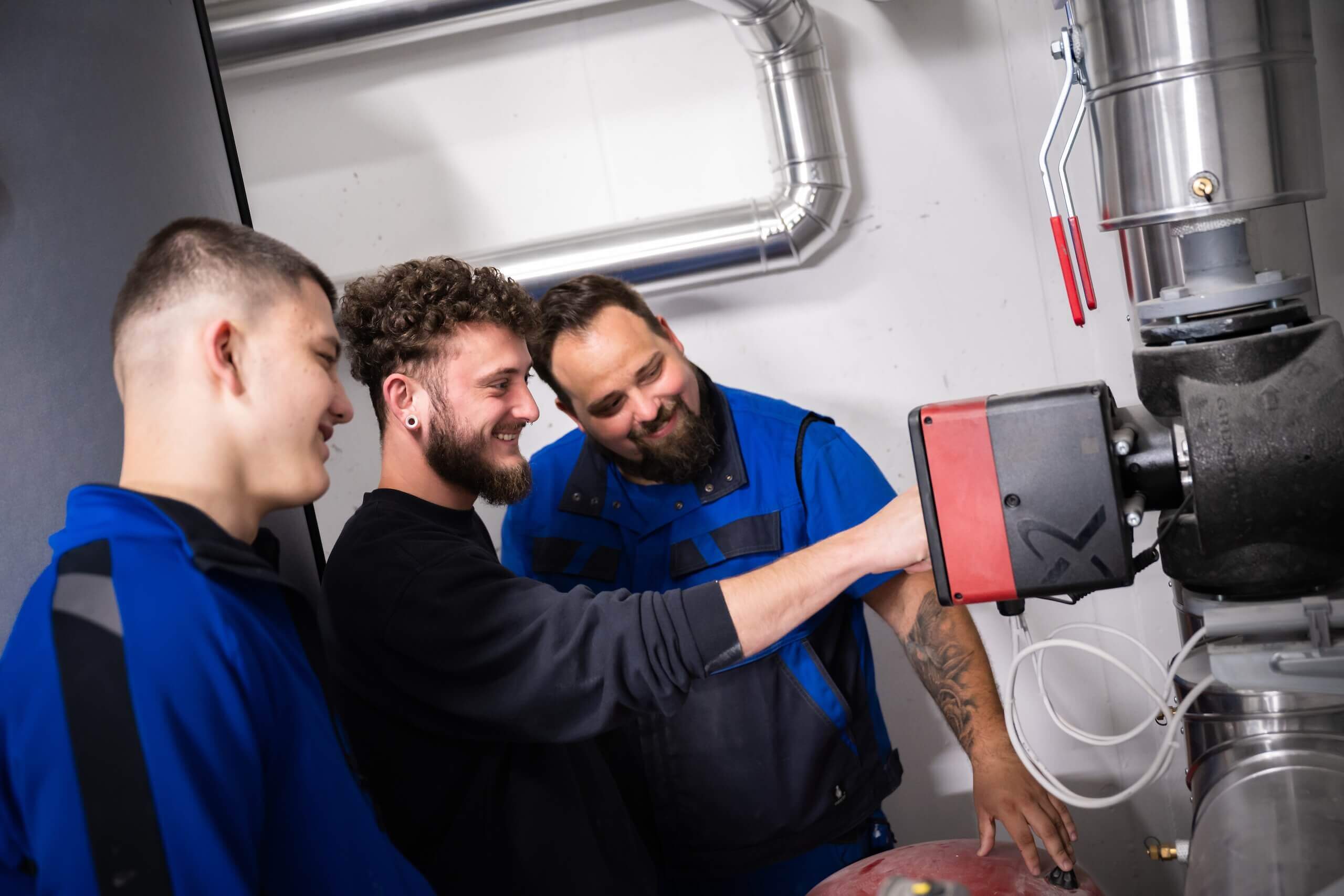 Drei Techniker arbeiten lächelnd an einer Heizungsanlage in einer industriellen Umgebung.