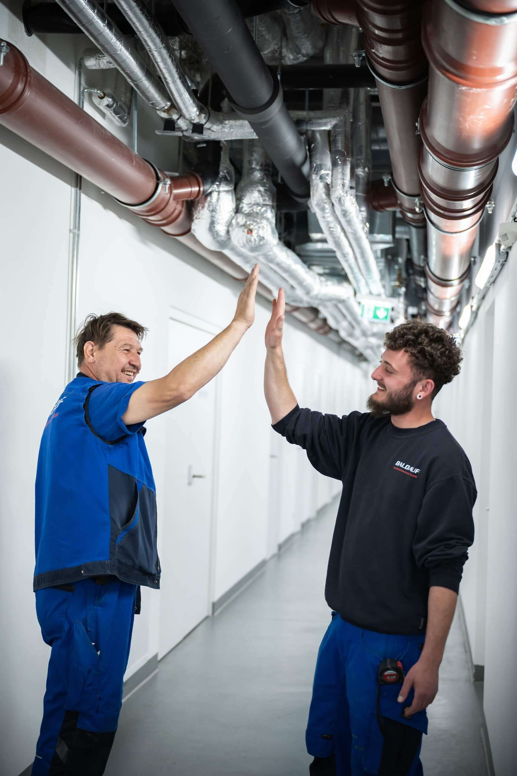 Zwei Handwerker in Arbeitskleidung geben sich in einem Gang unter Rohren ein High-Five.