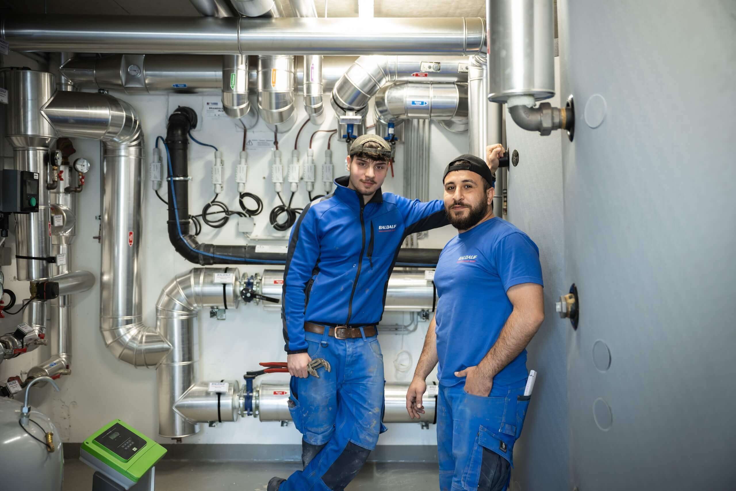 Installateure im Heizungsraum, umgeben von Rohren und Technik, in blauen Arbeitskleidung tätig.