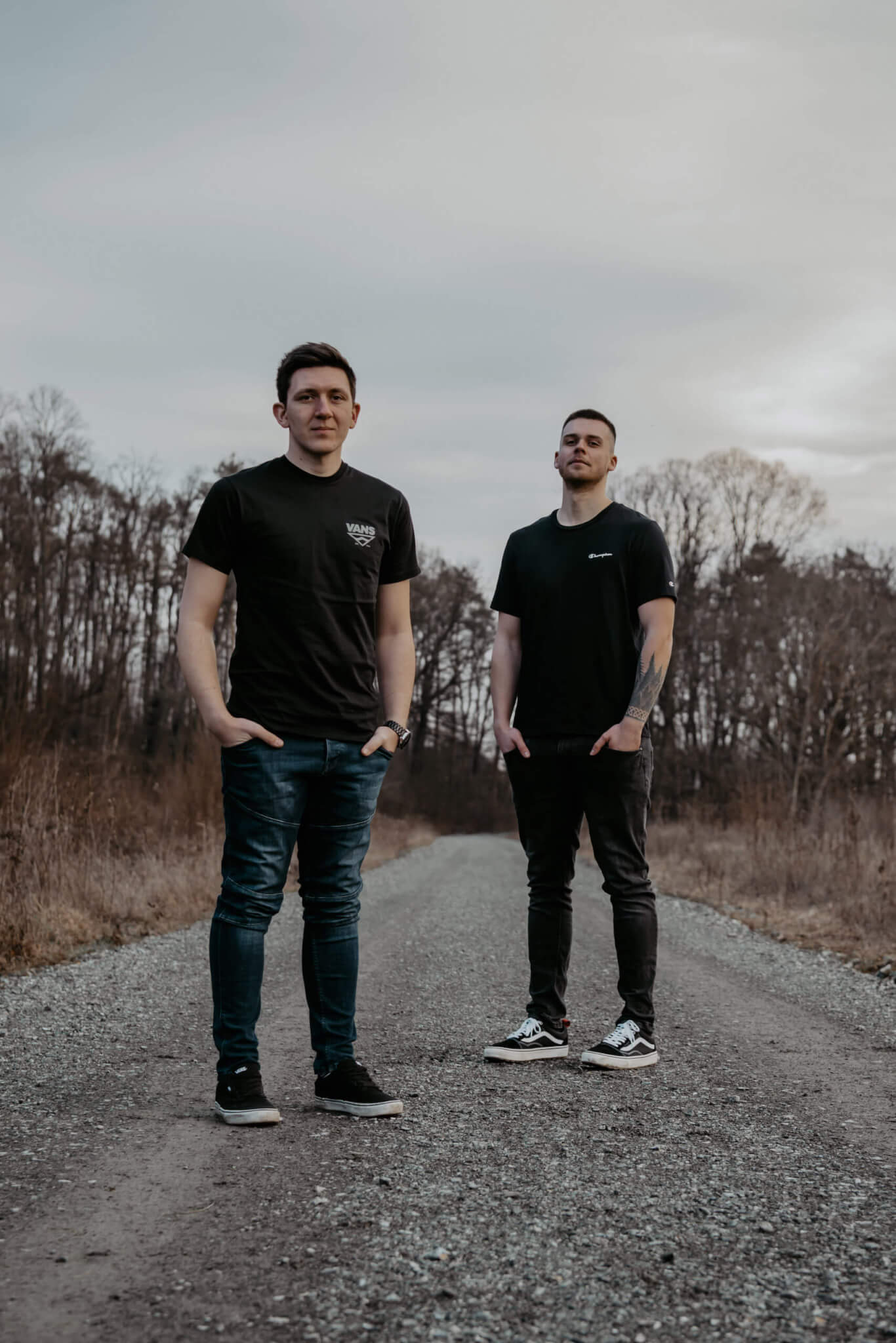 Zwei Männer in schwarzen T-Shirts stehen auf einem Schotterweg vor einem Wald, entspannte Outdoor-Szenerie.