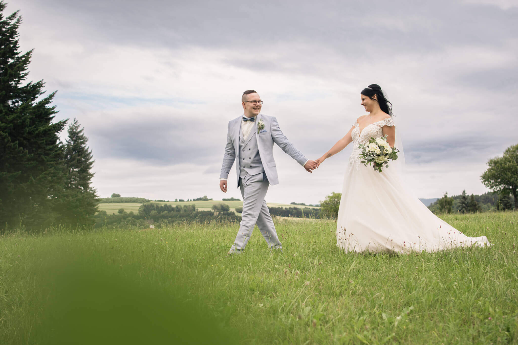 Brautpaar hält sich die Hände auf einer grünen Wiese, Hintergrund aus Wolken und Bäumen, romantische Hochzeitsaufnahme.