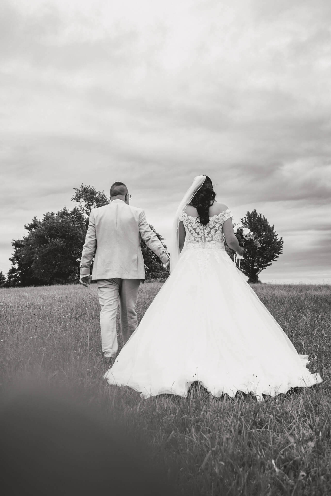 Brautpaar spaziert Hand in Hand über eine Wiese, Schwarz-Weiß-Hochzeitsfoto mit dramatischem Himmel.