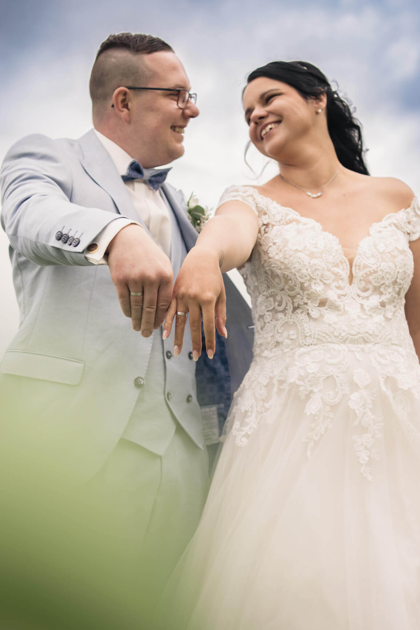 Hochzeitsfoto: Glückliches Brautpaar zeigt ihre Eheringe in eleganten Hochzeitsoutfits unter blauem Himmel.