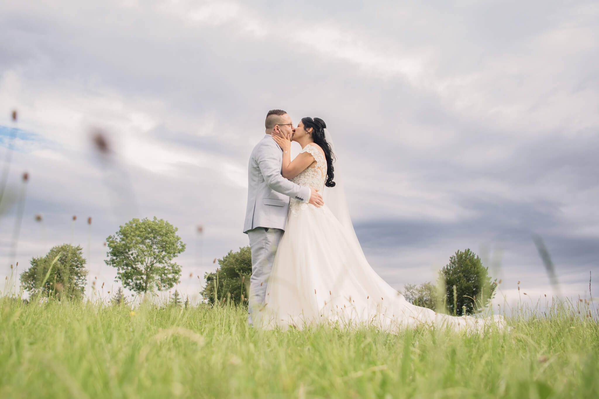 Brautpaar küsst sich auf grüner Wiese unter bewölktem Himmel an ihrem Hochzeitstag.