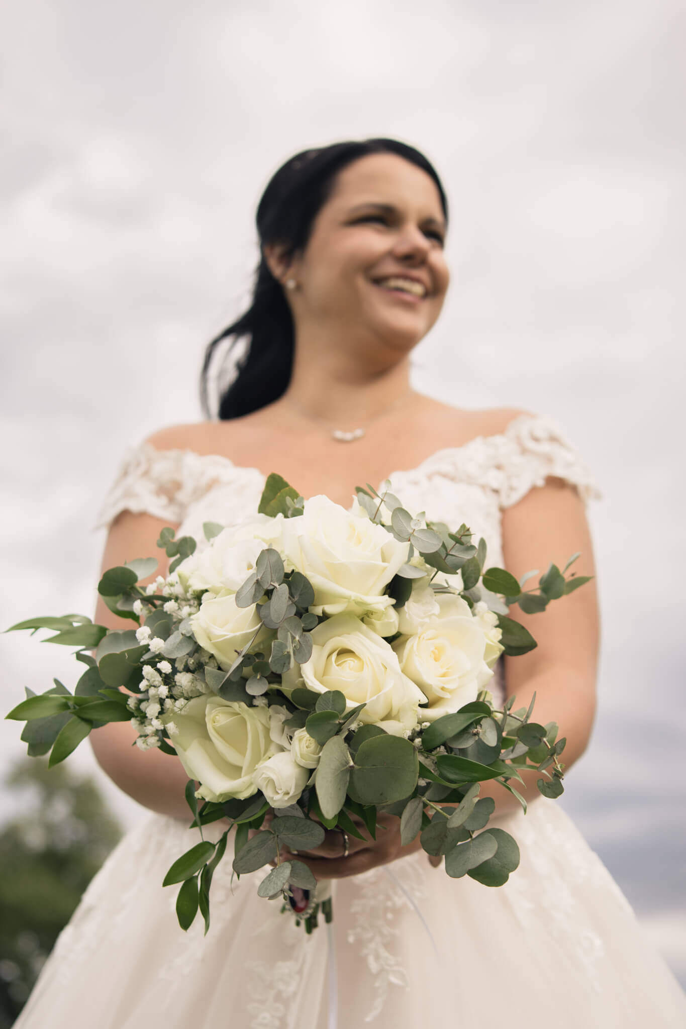 Braut im weißen Kleid hält eleganten Blumenstrauß aus weißen Rosen und Eukalyptus, himmelblauer Hintergrund.