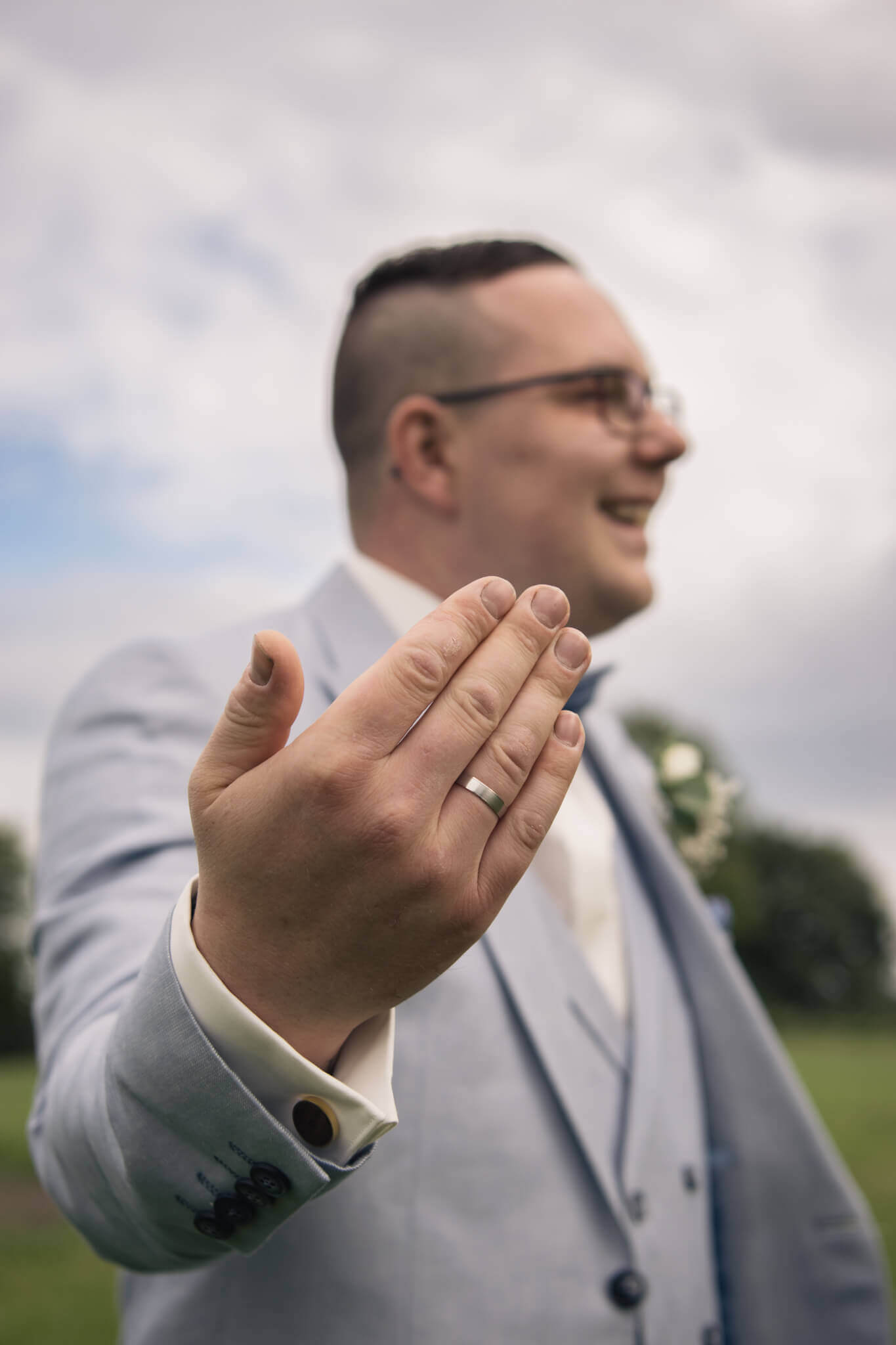 Lächelnder Bräutigam im grauen Anzug zeigt Ehering, glücklicher Moment bei sonniger Hochzeit im Freien.