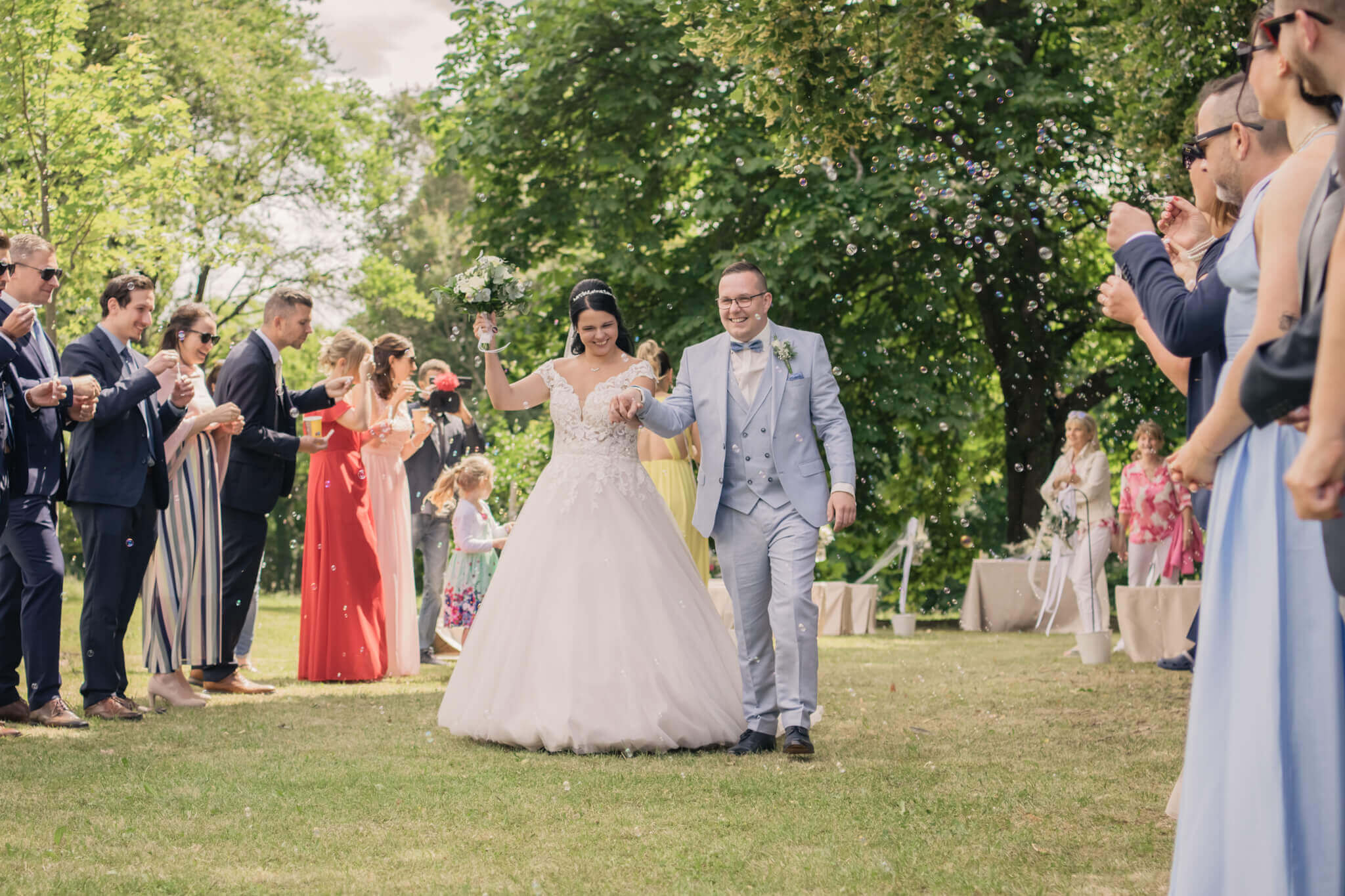 Braut und Bräutigam jubelnd bei einer Outdoor-Hochzeit, umgeben von Gästen und Seifenblasen im Grünen.