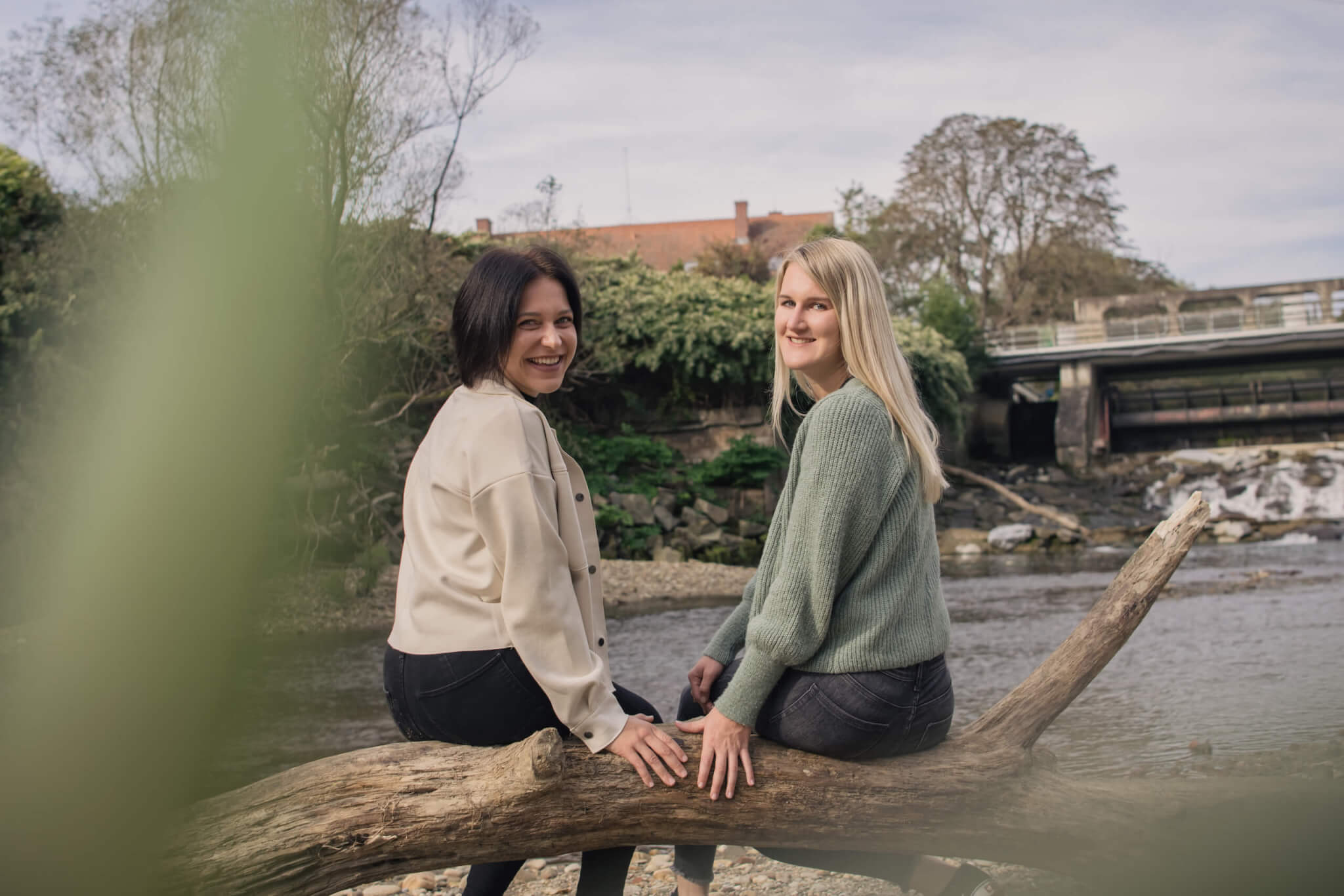 Zwei Frauen sitzen auf einem Baumstamm am Fluss, umgeben von Natur, unterhalten sich und genießen den Ausblick.