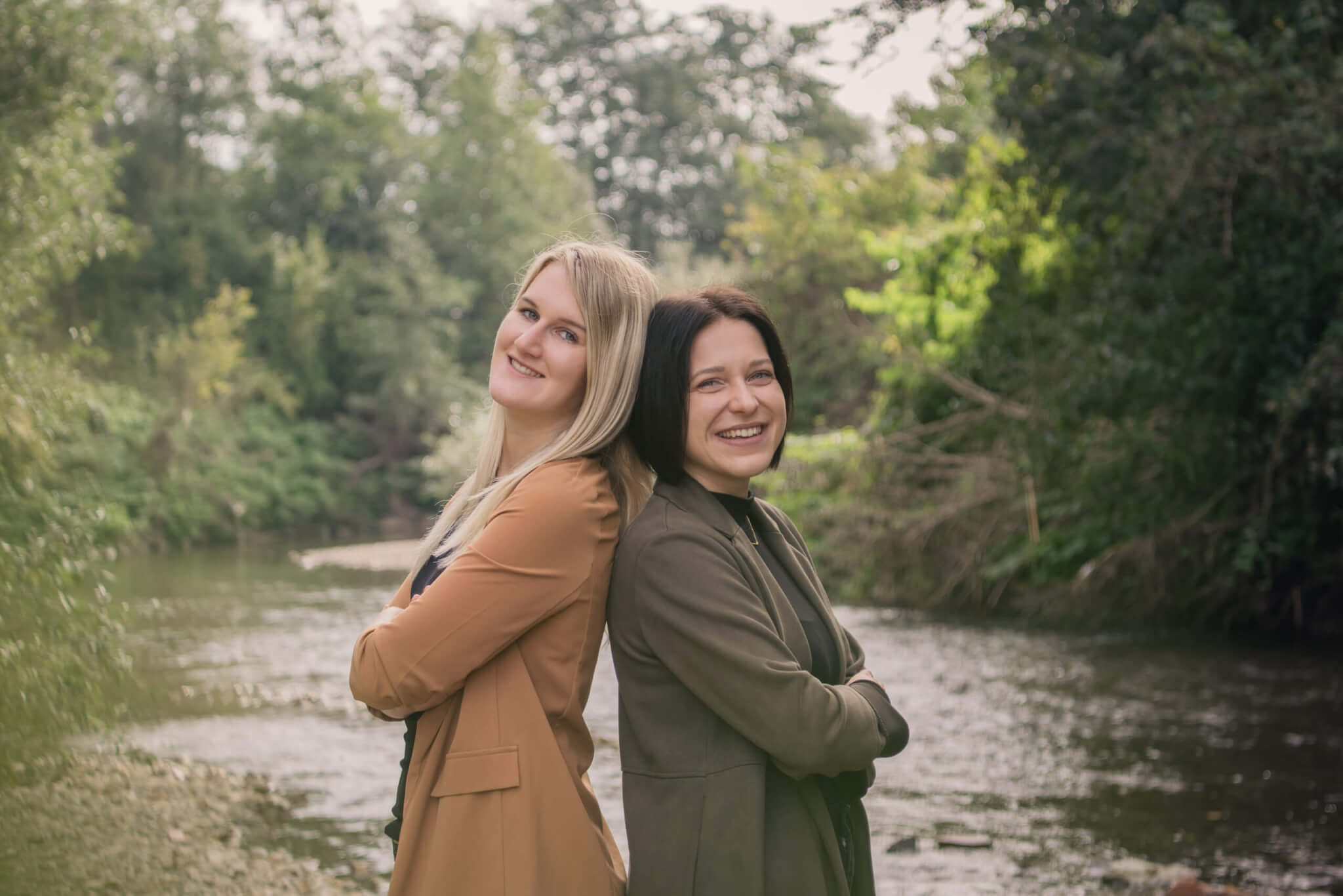 Zwei Frauen stehen Rücken an Rücken und lächeln in der Natur am Flussufer.