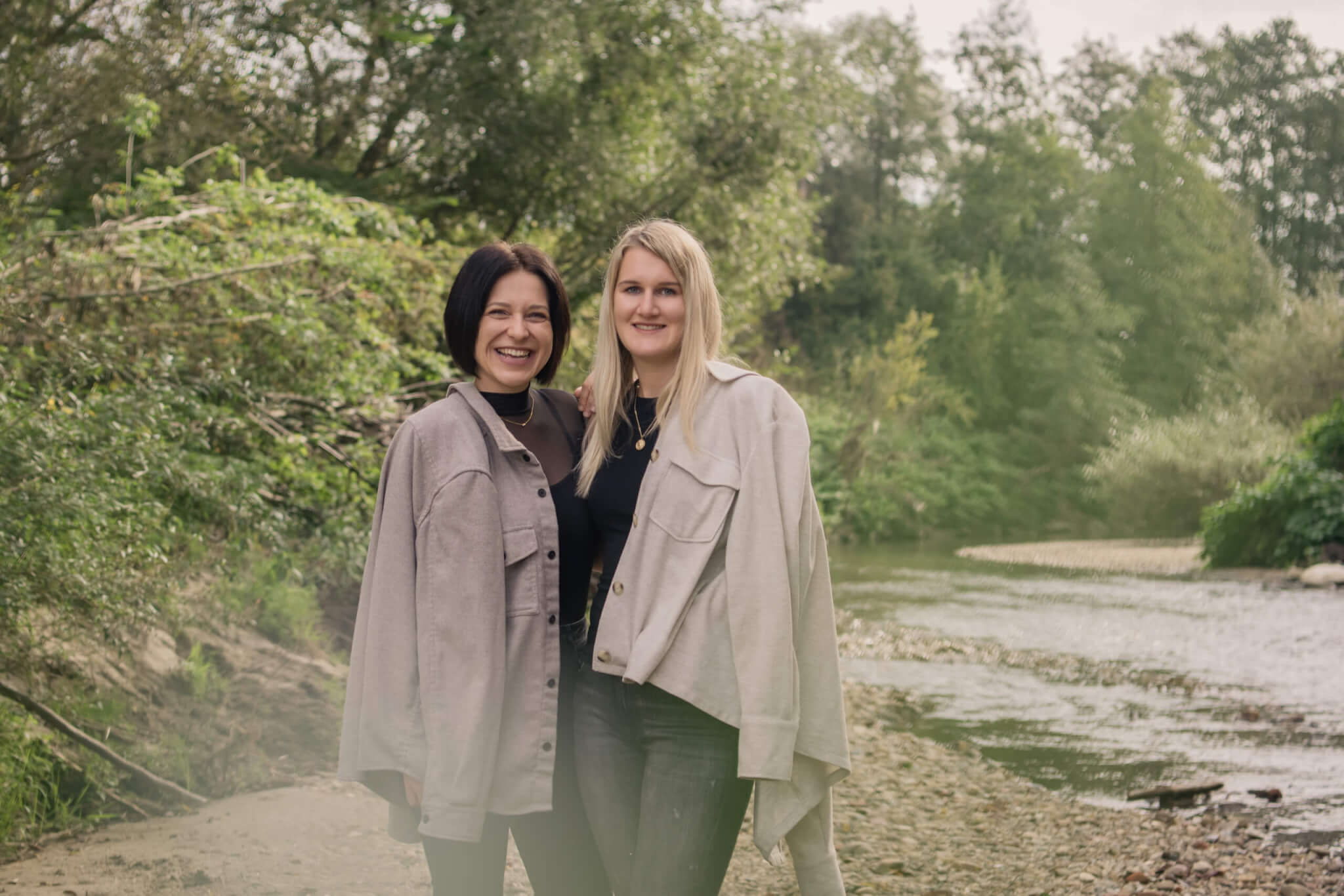 Zwei Frauen lächeln im Grünen am Flussufer, tragen helle Jacken, Natur im Hintergrund.