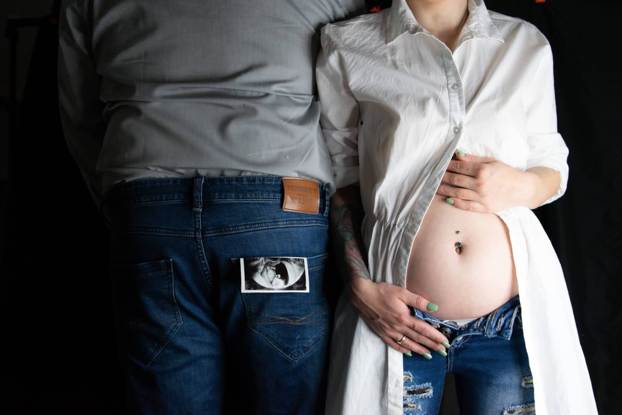 Paar zeigt Schwangerschaftsstolz: Ultraschallbild in Jeanstasche, Hand auf Babybauch in offener Bluse.