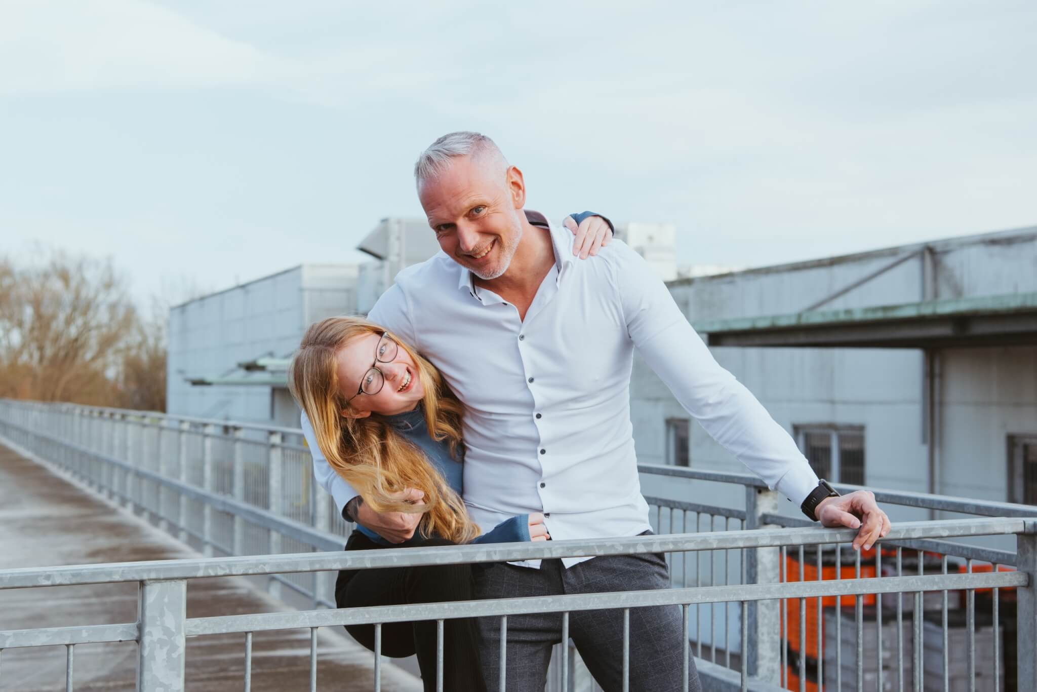 Vater und Tochter lachend auf einer Brücke, beide in heller Kleidung, genießen die gemeinsame Zeit im Freien.