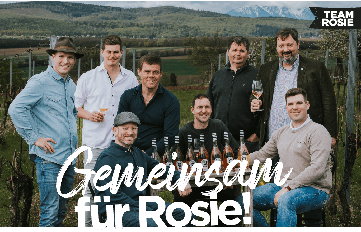 Gruppe von acht Männern in einem Weinberg hält Weinflaschen, mit dem Schriftzug Gemeinsam für Rosie.