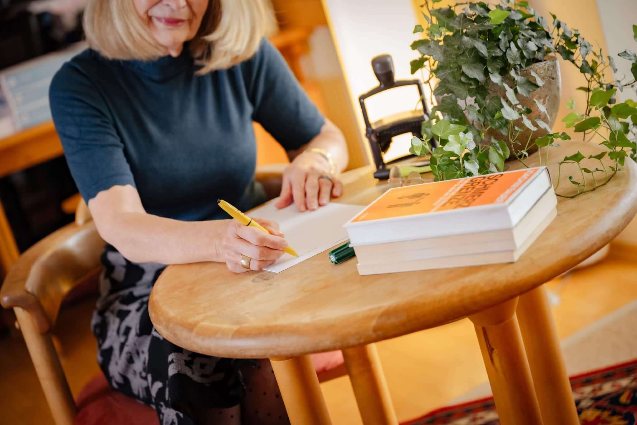 Frau schreibt Notizen am Holztisch mit Buch und Pflanze, idealer Ort für kreative Arbeiten und Inspiration.