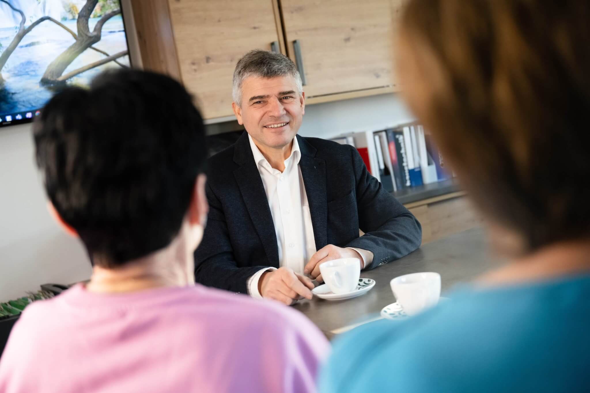 Mann im Anzug bei Besprechung mit Kaffee, lächelt zwei Personen an. Bürohintergrund mit Bildern und Akten.