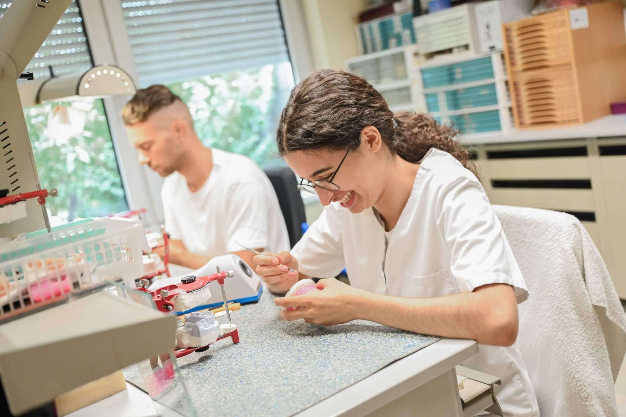 Zahntechnikerin arbeitet konzentriert mit Dentalwerkzeugen am Laborarbeitsplatz, während ein Kollege im Hintergrund arbeitet.