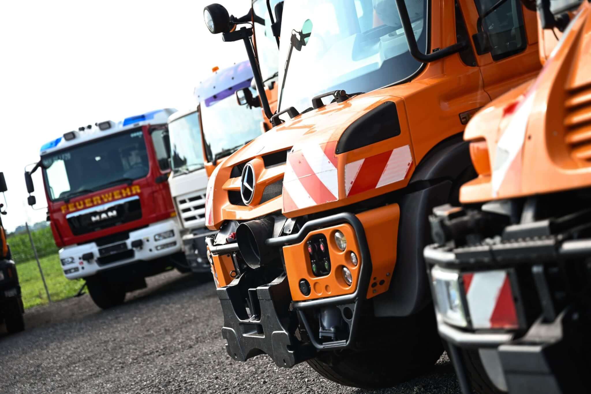 Reihe von Einsatzfahrzeugen, darunter Feuerwehr- und orangefarbene Fahrzeuge auf einem Schotterplatz.