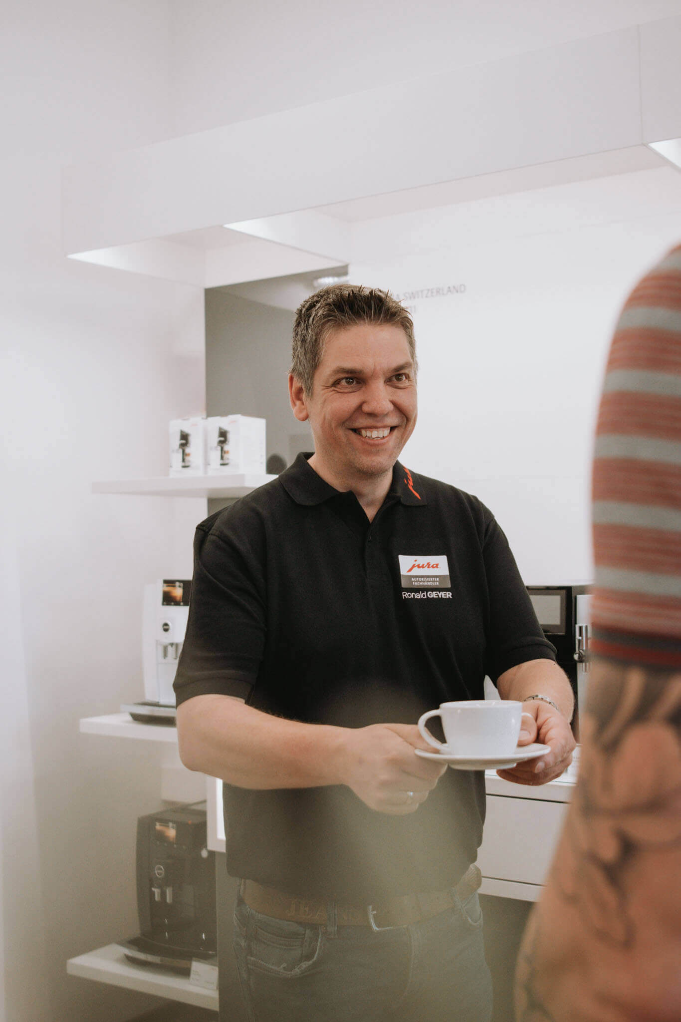 Person serviert Kaffee in einem modernen Geschäft, lächelt freundlich. Gerätekulisse im Hintergrund.